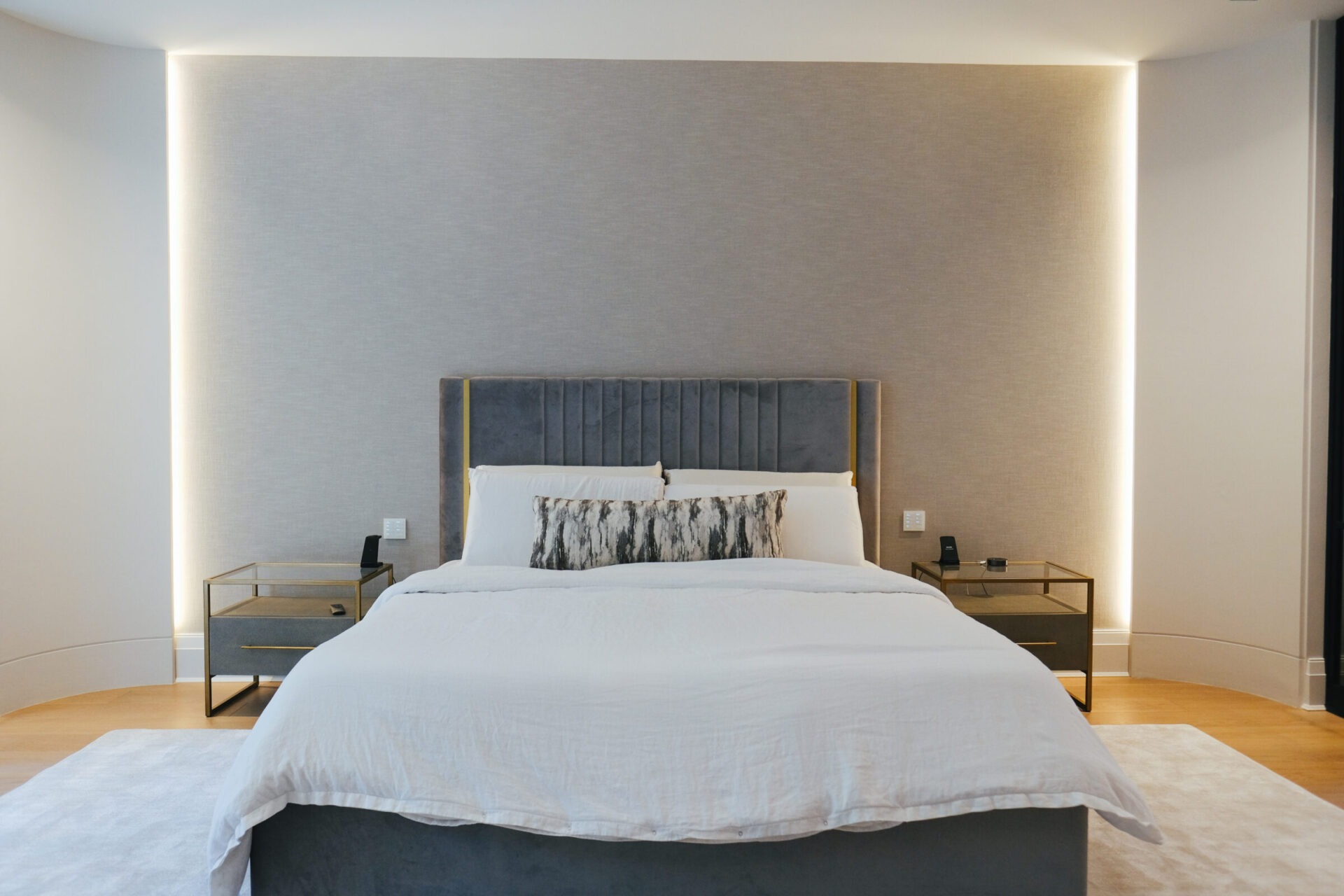 A modern bedroom with a sleek, gray upholstered bed, soft lighting, and minimalist bedside tables on a wooden floor with a white rug.