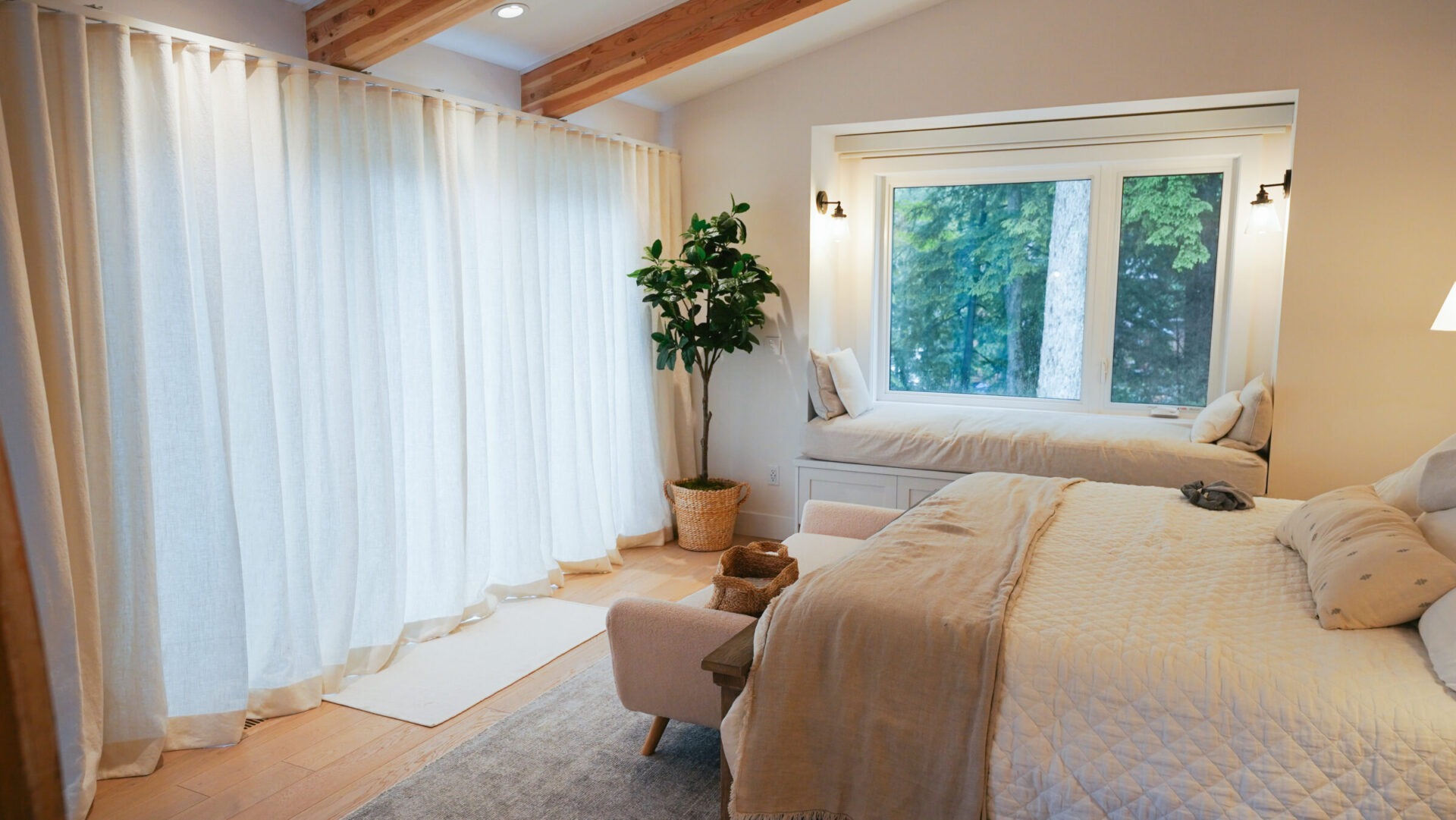 A cozy bedroom with large windows, a comfortable bed, neutral décor, and a potted plant. Soft lighting creates a serene atmosphere.