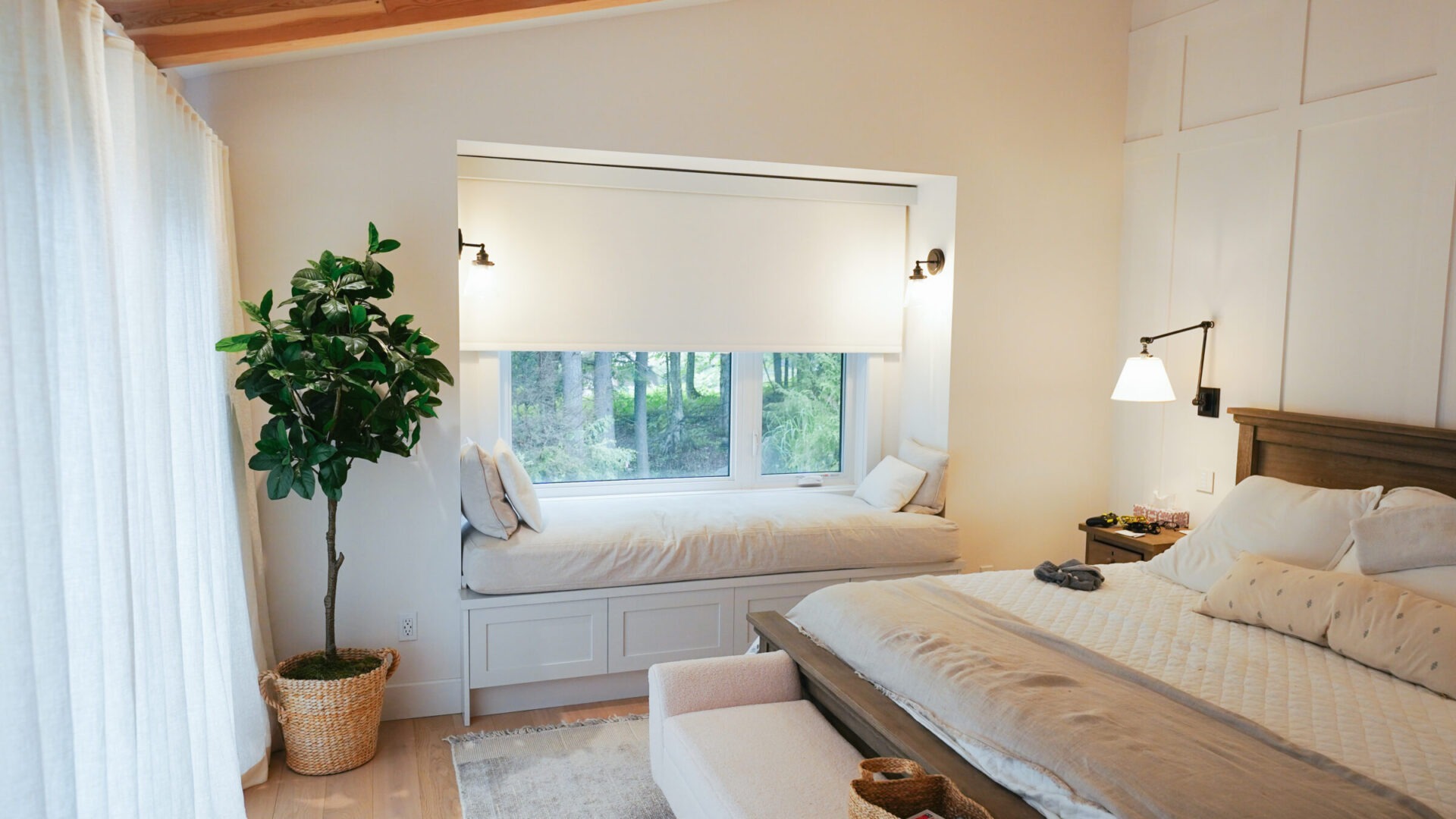 Cozy bedroom with neutral tones, a large bed, window seat, and indoor plant. Sunlight filters through sheer curtains, creating a peaceful atmosphere.