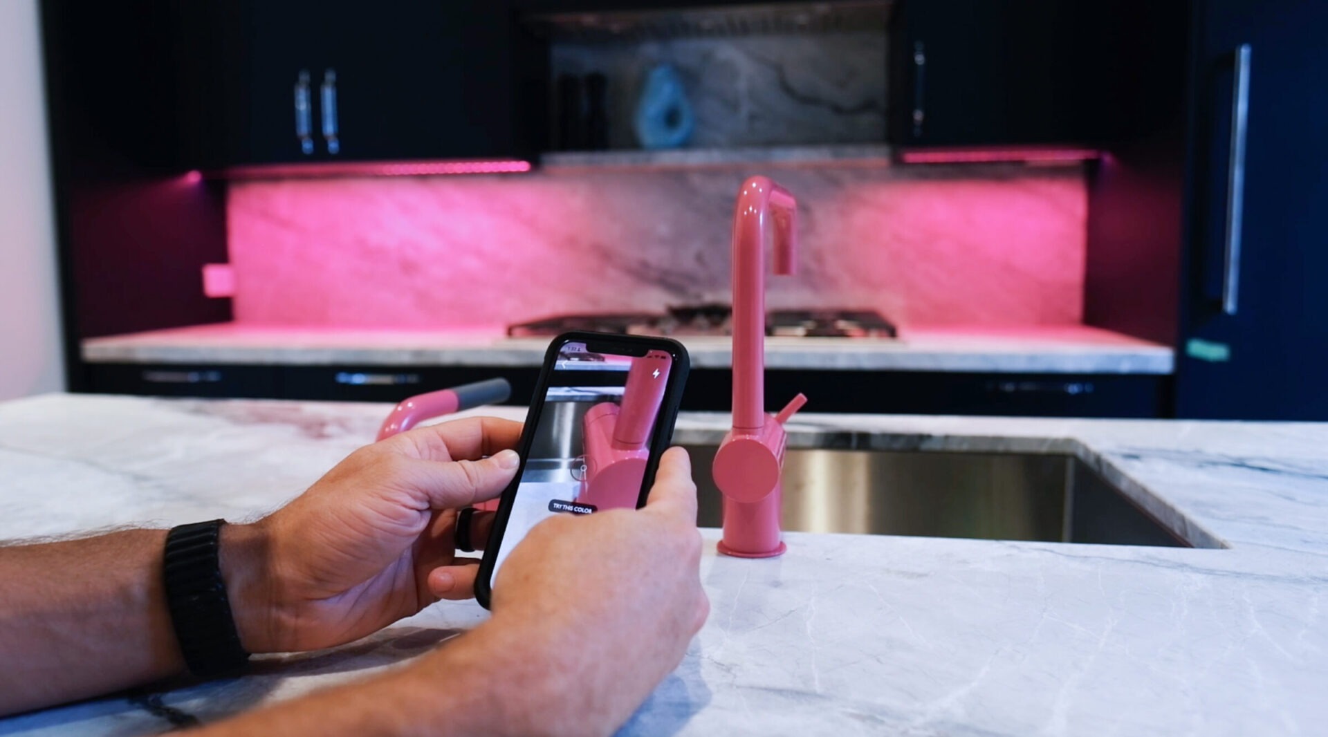 A person controls a pink kitchen faucet using a smartphone app. The kitchen has a sleek marble countertop and pink LED lighting.