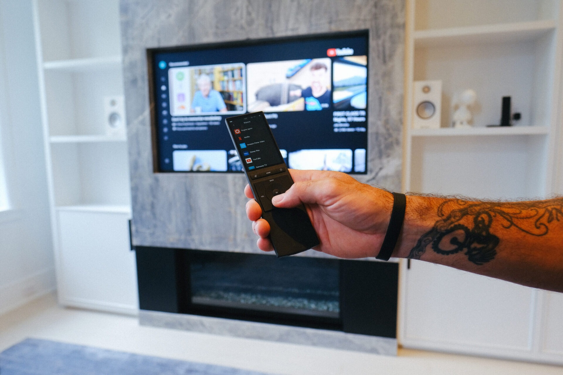 A person holds a remote control, pointing towards a wall-mounted TV displaying a video streaming platform. Modern living room setting with speakers.