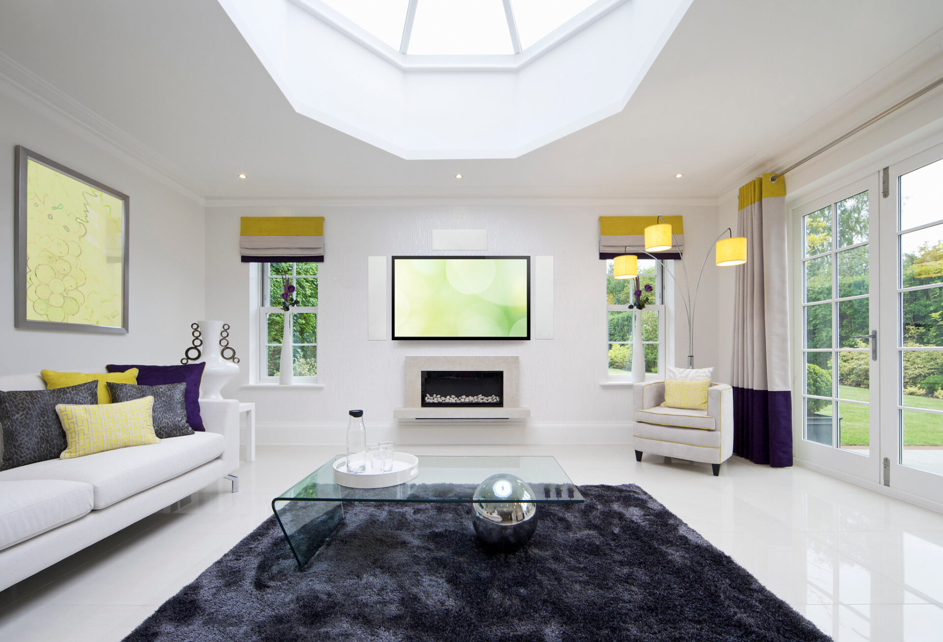 A modern living room with a skylight, white furniture, abstract art, and a glass coffee table. Large windows overlook a garden.