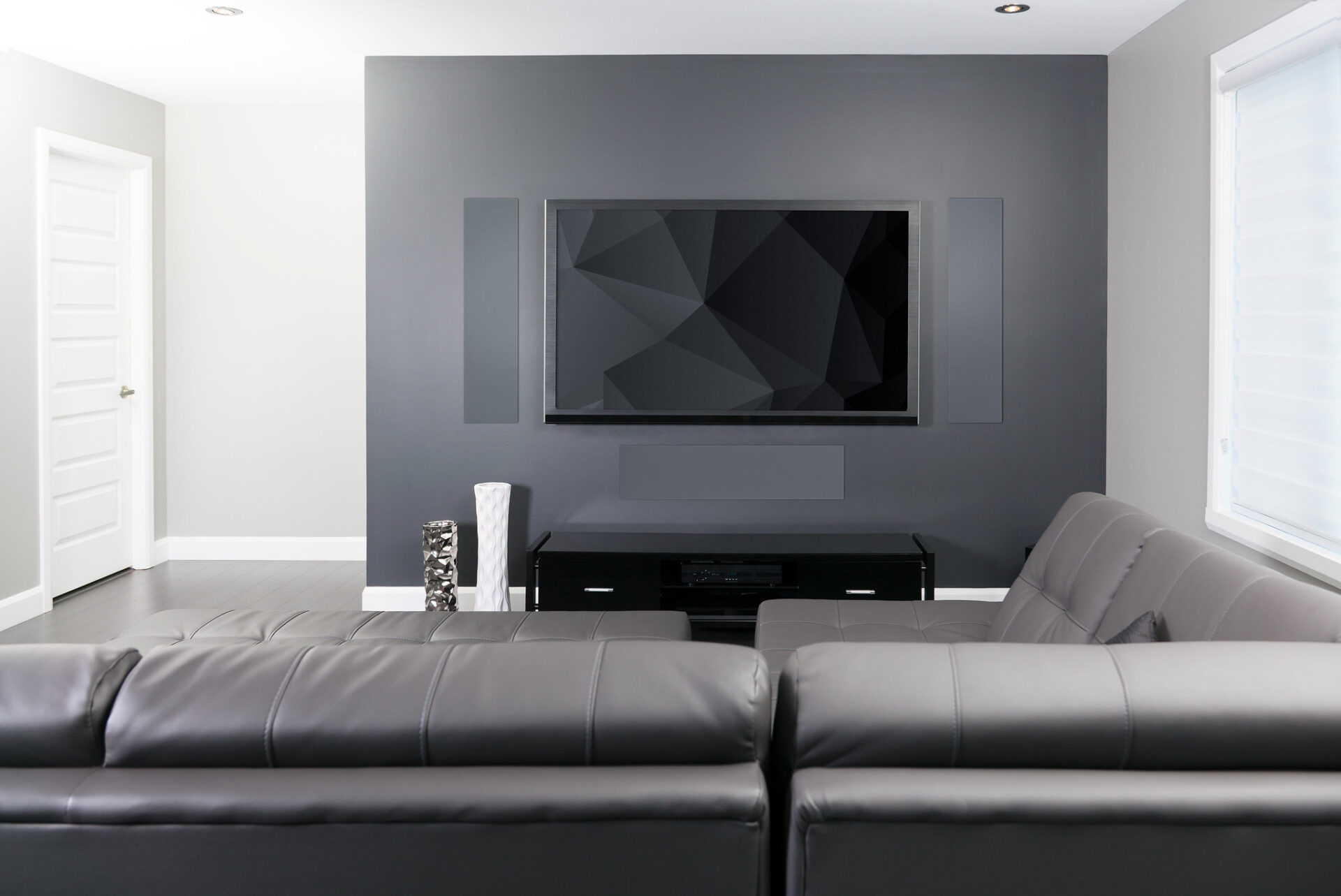 Modern living room with a large flat-screen TV, two grey couches, white walls, and minimalist decor, including a geometric wall panel design.