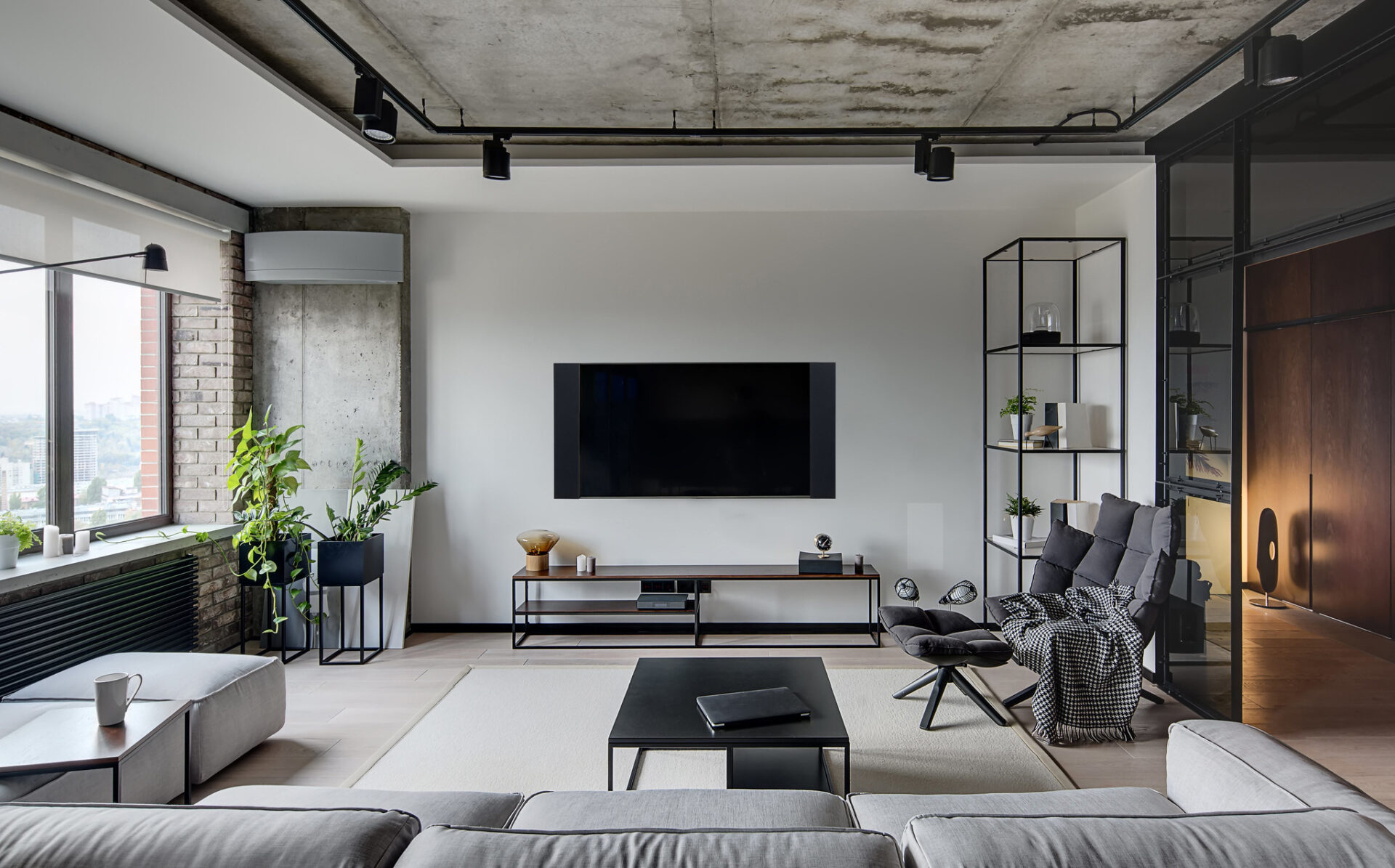 Modern living room with minimalist decor, featuring a large TV, sofa, coffee table, and indoor plants. Bright, spacious ambiance with industrial elements.