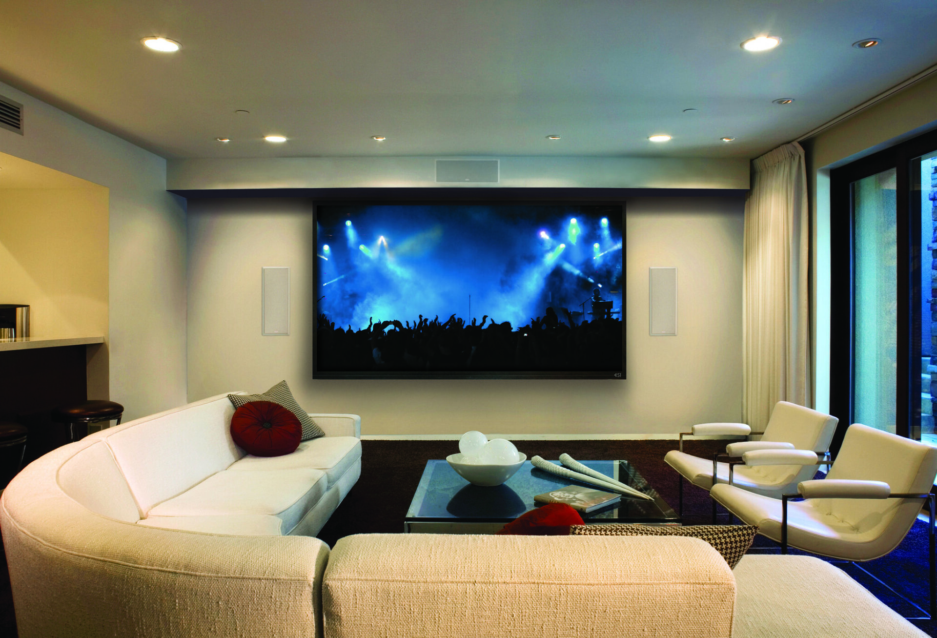 Modern living room with a large screen displaying a concert. White sofas surround a glass coffee table. Soft lighting and floor-to-ceiling windows.