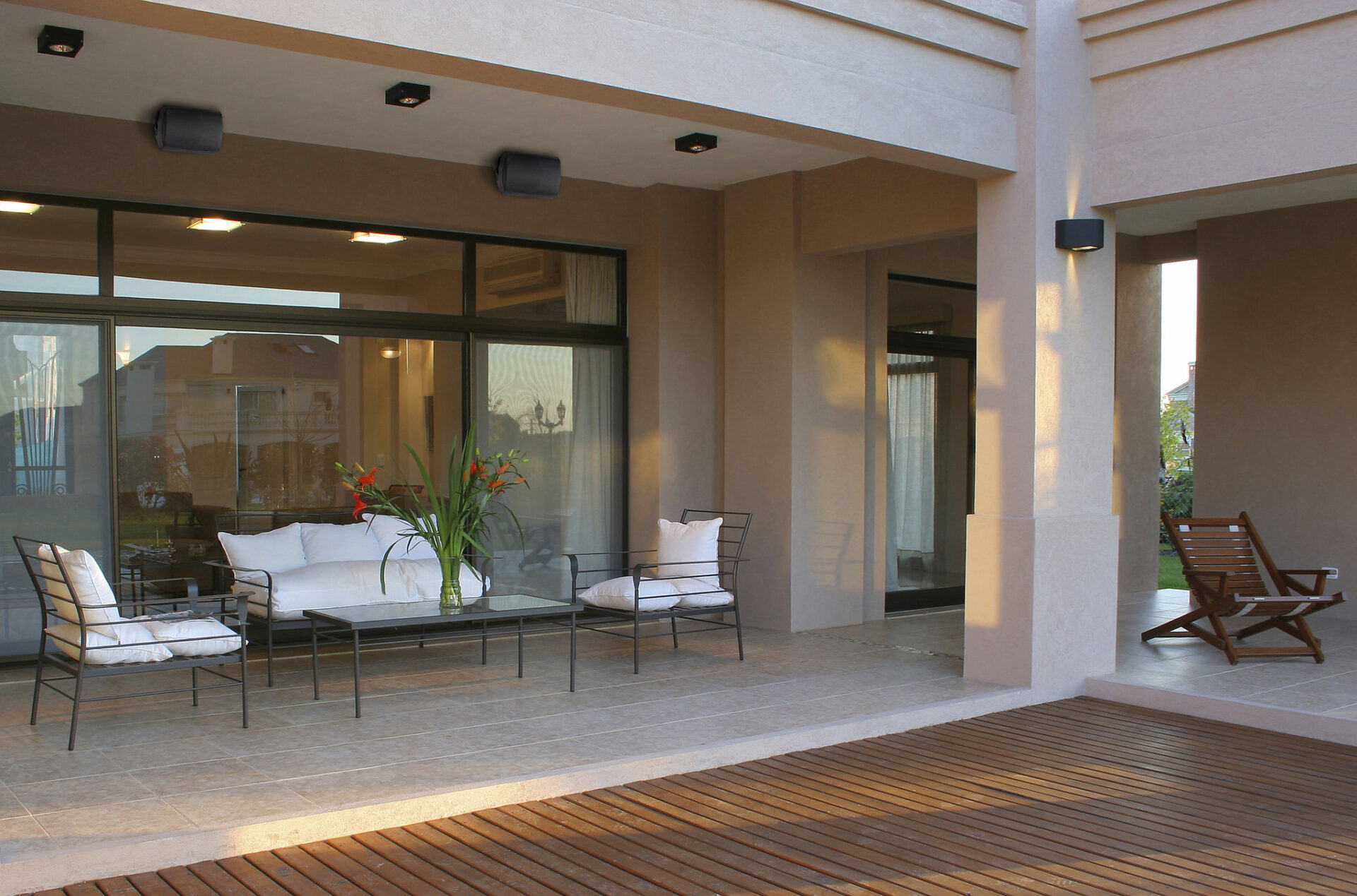 Modern patio with cushioned seating and glass doors, surrounded by beige columns. Wooden deck extends from outdoor living space, adorned with potted plants.