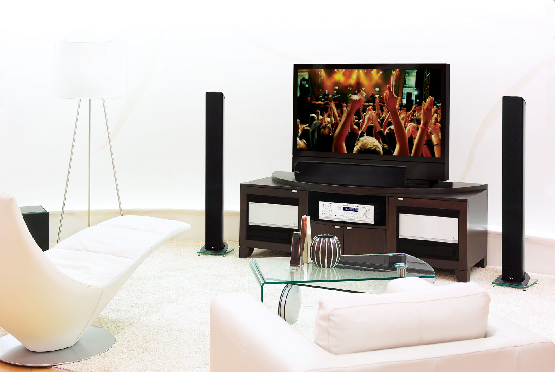 A modern living room features a flat-screen TV showing a concert, sleek white furniture, a glass coffee table, and tall speakers.