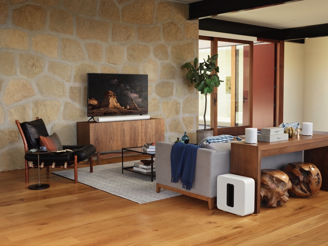 A modern living room with wooden furniture, large stone wall, and television. Stylish decor includes plants, cushions, and abstract sculptures on natural wood floor.