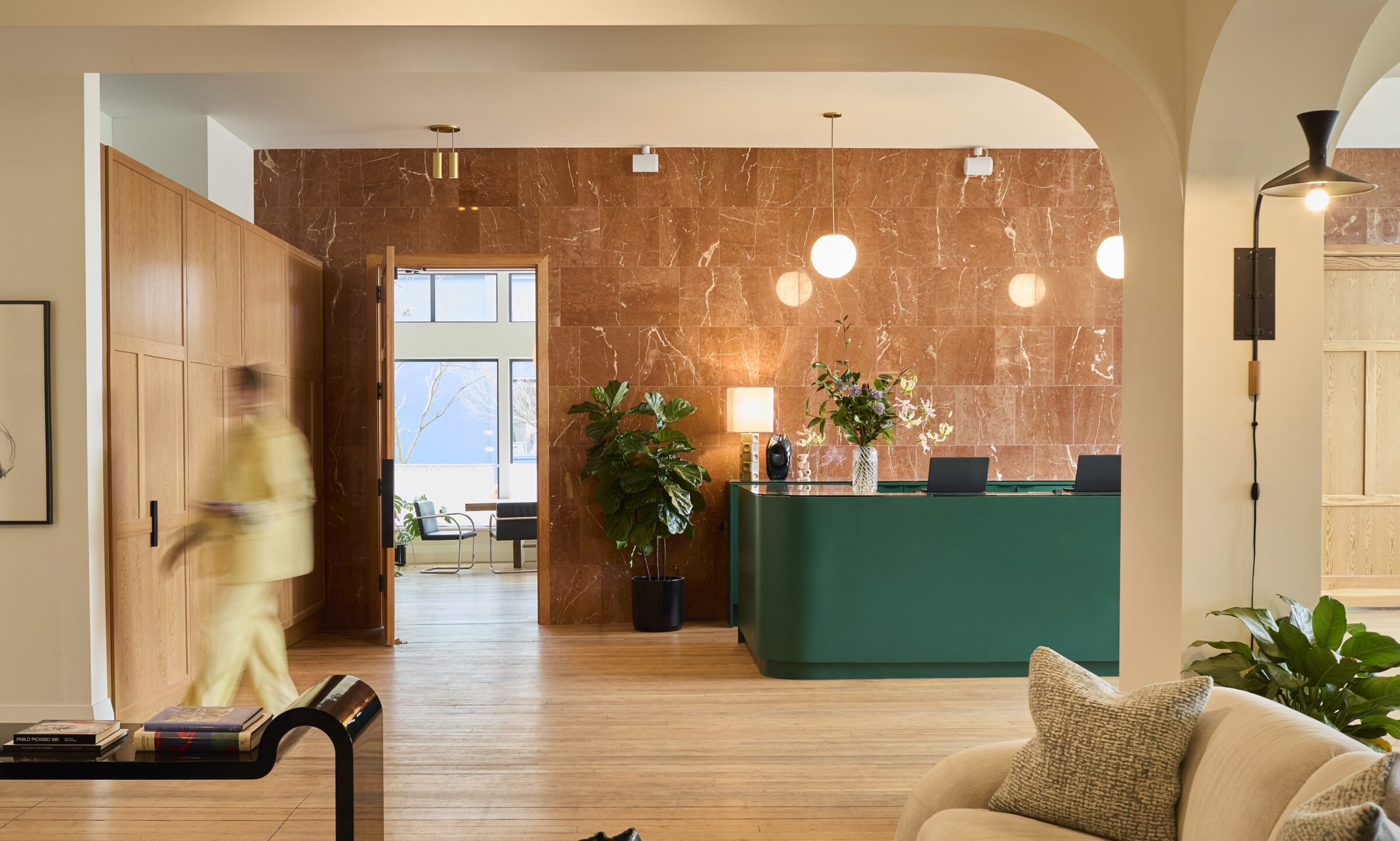 A modern lobby with green reception, wooden accents, and stylish lighting. A person walks past, with plants and seating enhancing the cozy atmosphere.