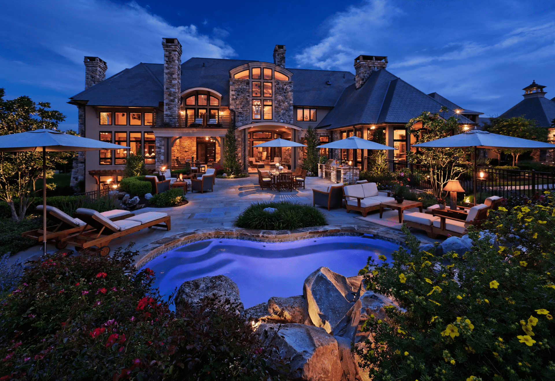 Luxurious stone mansion at twilight, featuring a landscaped patio, glowing pool, seating areas, and soft ambient lighting amid lush gardens.