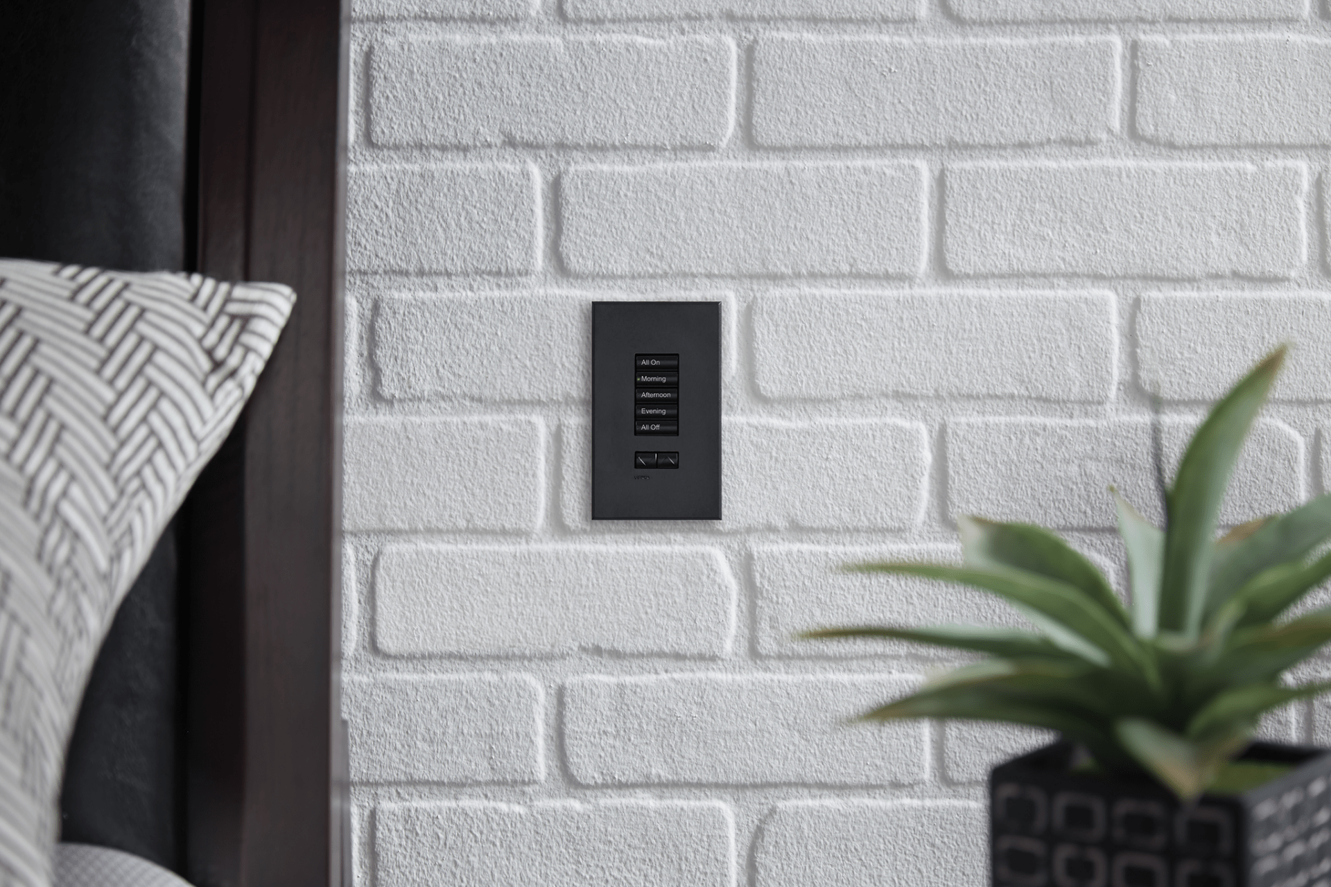 A white brick interior wall with a black light switch, next to a patterned pillow and a potted plant on a table.