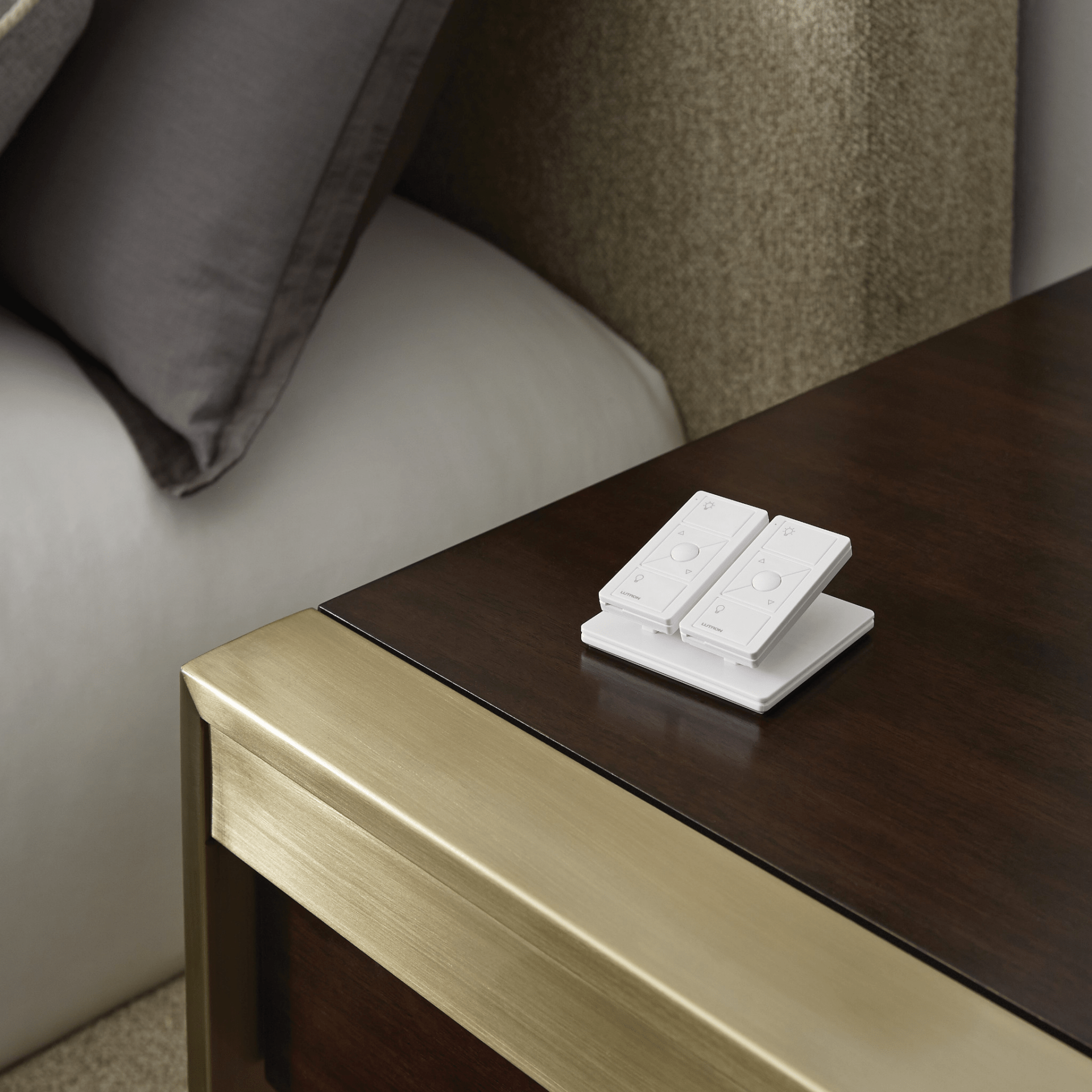 A wooden and metallic nightstand holds a small stack of white remote controls beside a neatly made bed with grey and white pillows.