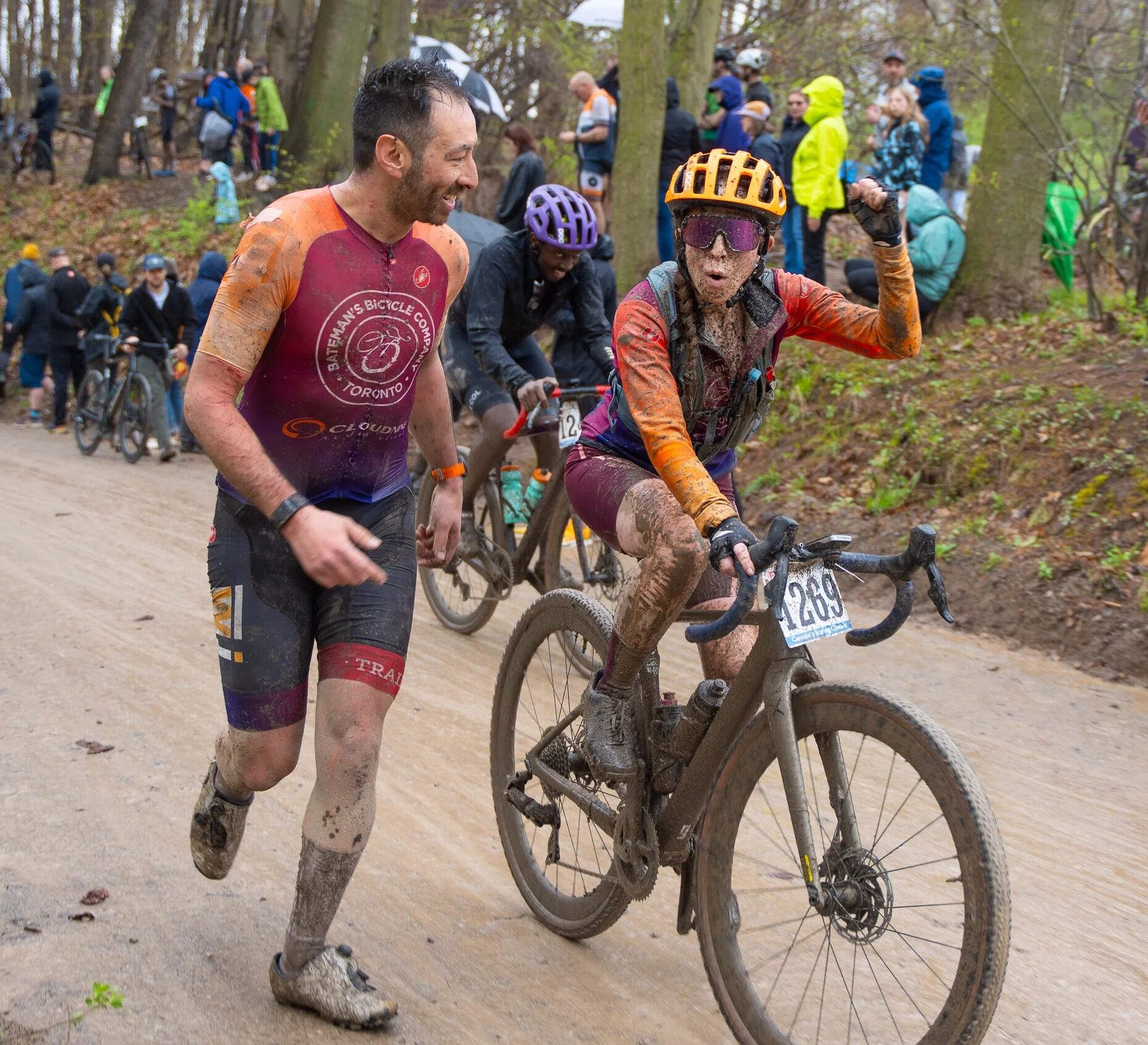 Cyclist and person celebrate on a muddy trail during an outdoor biking event, surrounded by cheering spectators in a wooded area.