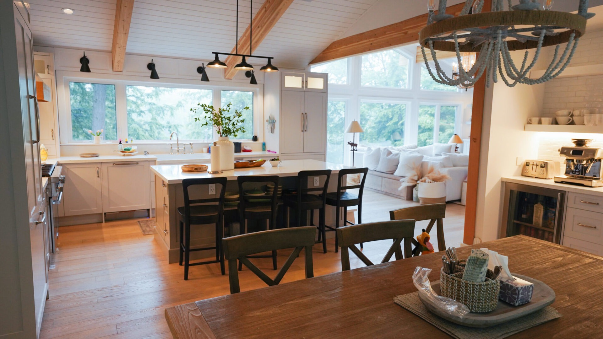 Spacious kitchen and living area with wooden beams, large windows, and cozy seating. Features modern appliances, island with stools, and decorative chandelier.