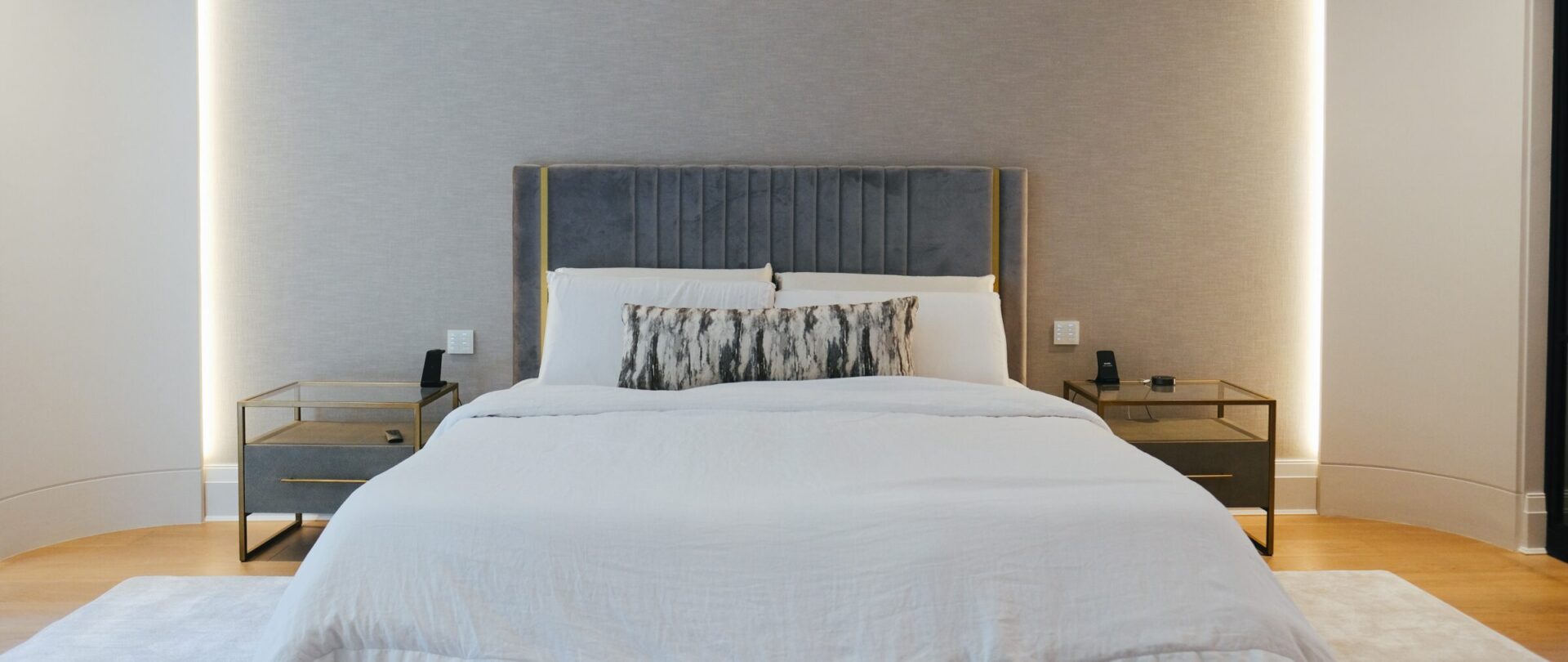 Modern bedroom with a neatly made bed featuring a grey headboard, flanked by two bedside tables. Subtle lighting creates a cozy ambiance.