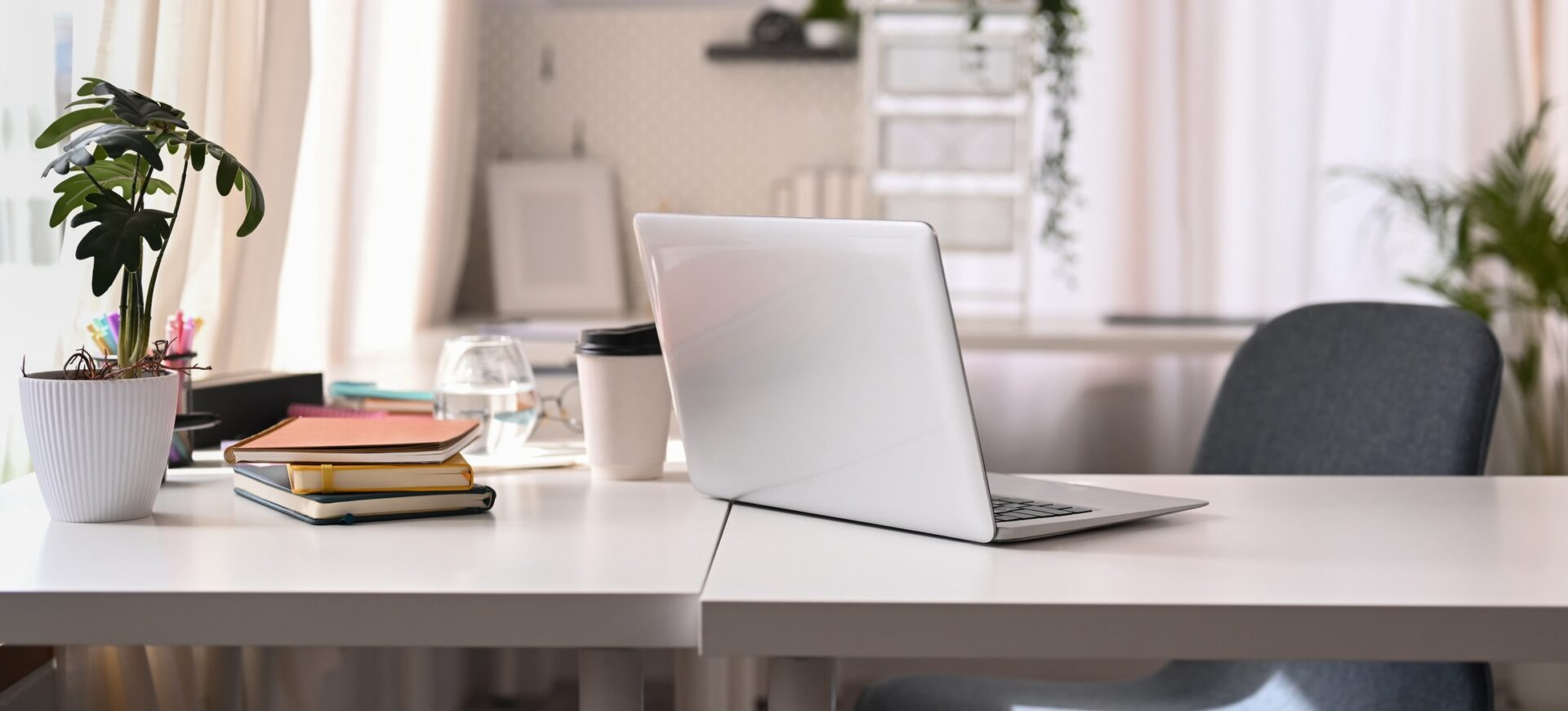 A minimalist home office with a laptop, coffee cup, books, and a plant on a white desk, near a chair and bright window.