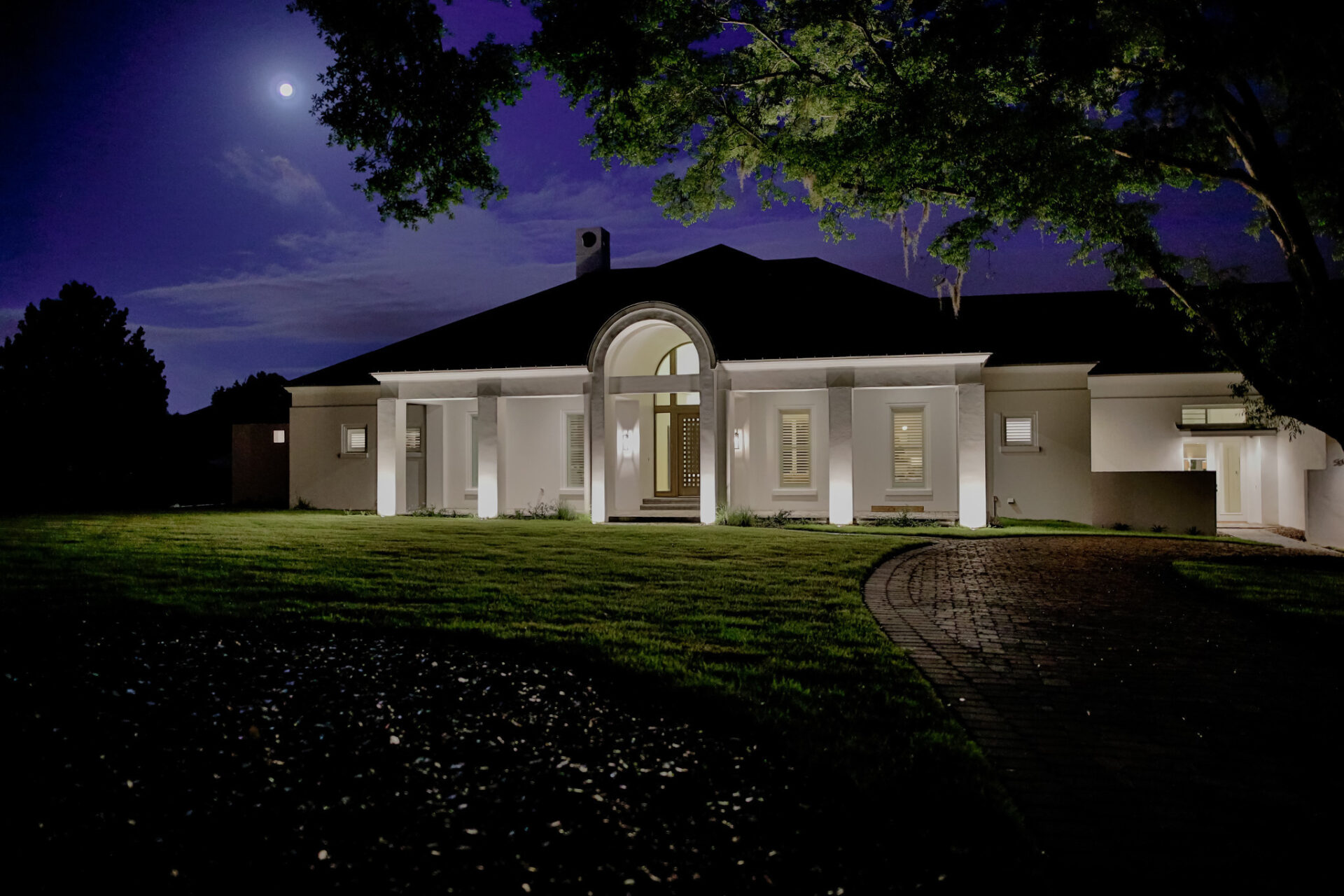 Elegant house illuminated at night, featuring arched entrance, manicured lawn, and a moonlit sky. Serene atmosphere with surrounding trees and pathway.