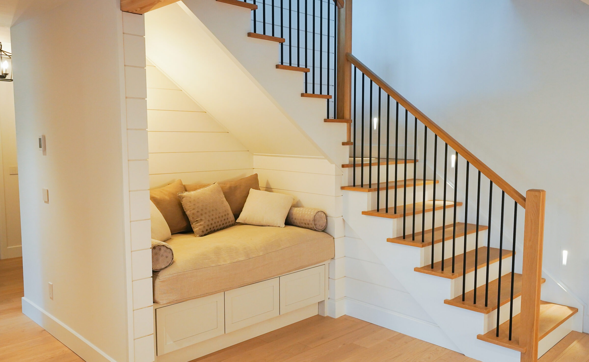 A cozy nook with cushions is located under a wooden staircase. The room features minimalistic design with white walls and a warm atmosphere.