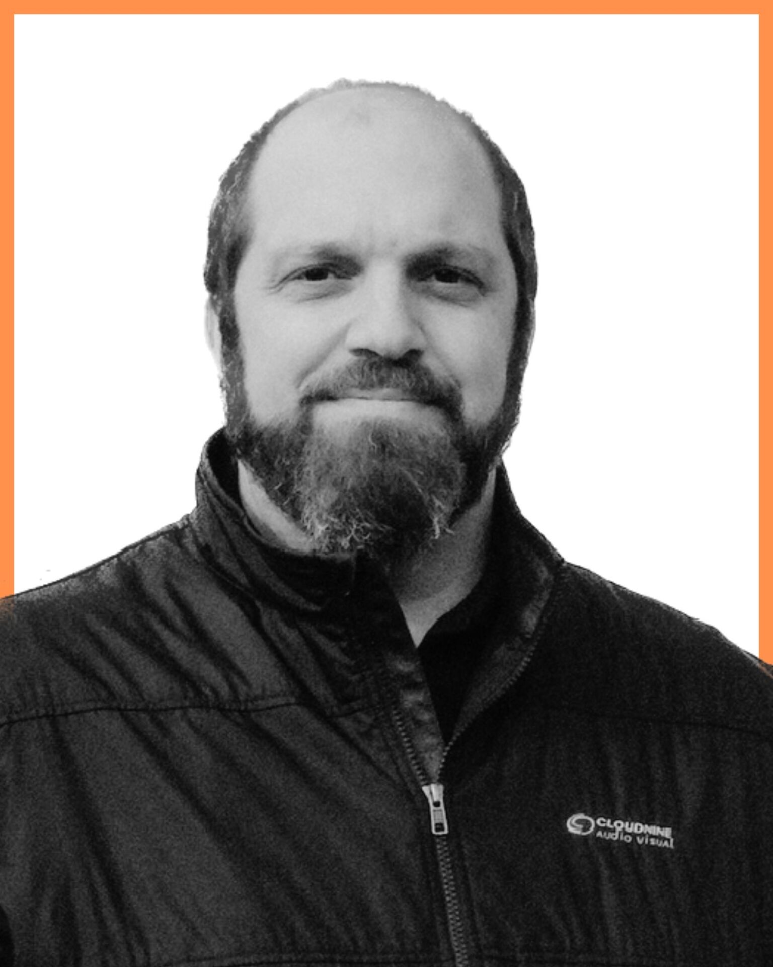 A person with a beard, wearing a jacket with a logo, is smiling against a plain white background bordered by an orange frame.