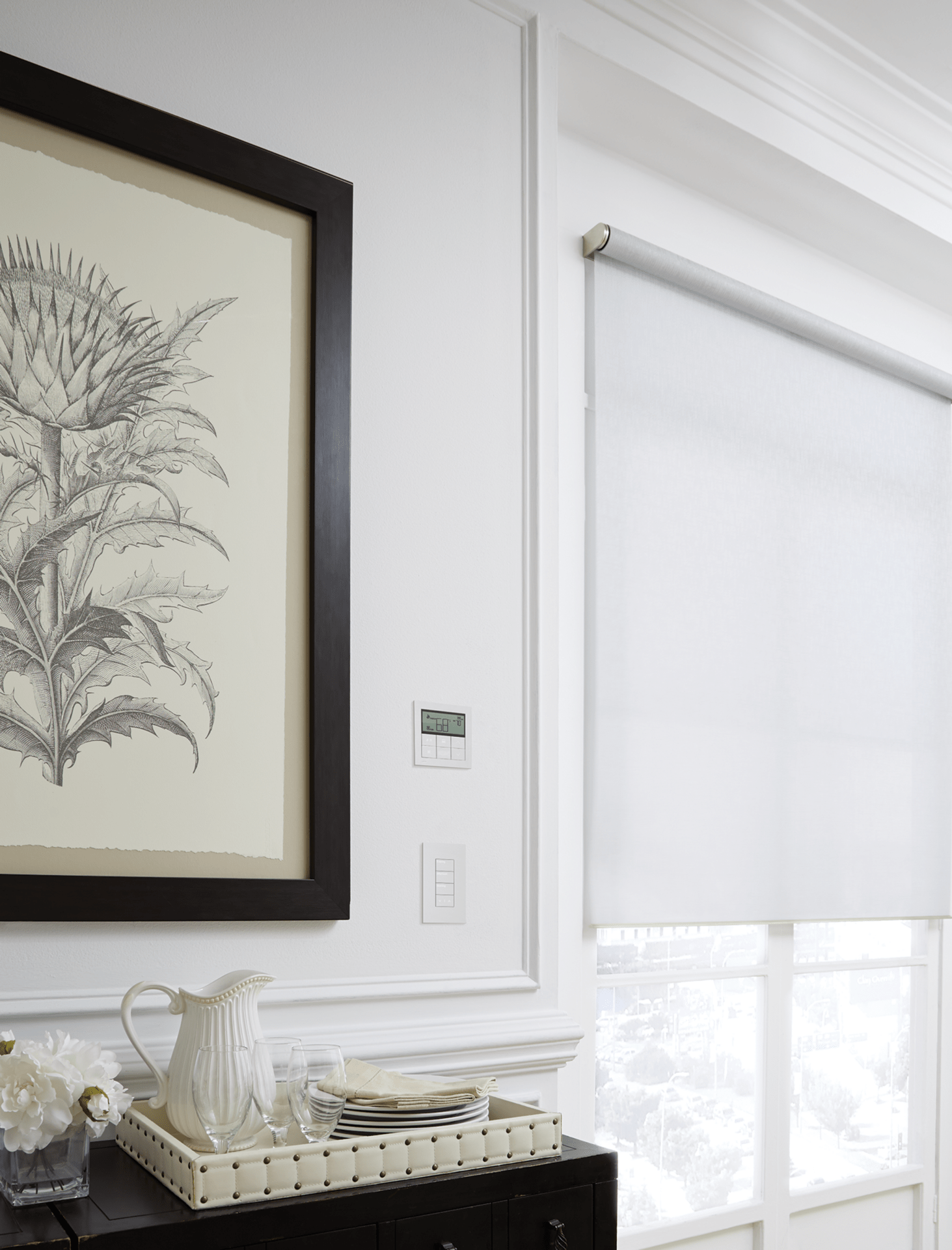 A minimalist interior with a framed botanical print, white roller blinds, and a tray with a pitcher and plates on a dark cabinet.