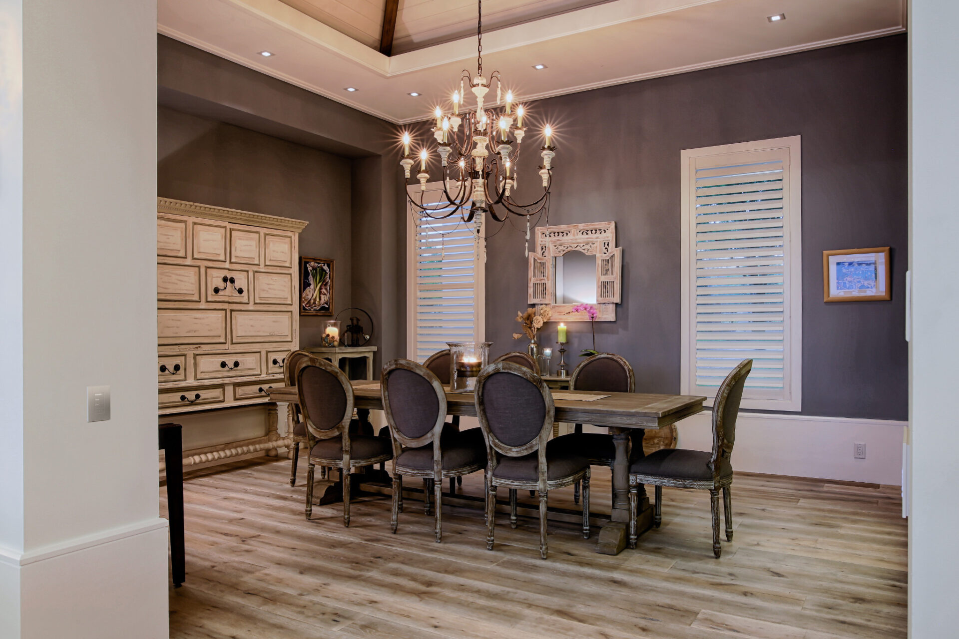 Elegant dining room with wooden table, eight upholstered chairs, ornate chandelier, and decorative cabinets. Cozy atmosphere enhanced by soft lighting and dark walls.