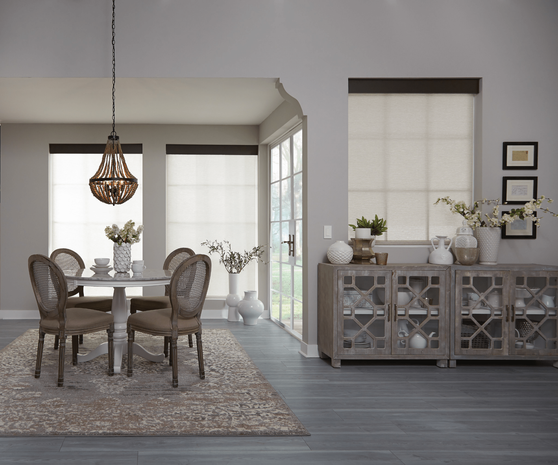 A modern dining room features a round table, four chairs, pendant light, sideboard, decorative vases, and large windows with neutral shades.