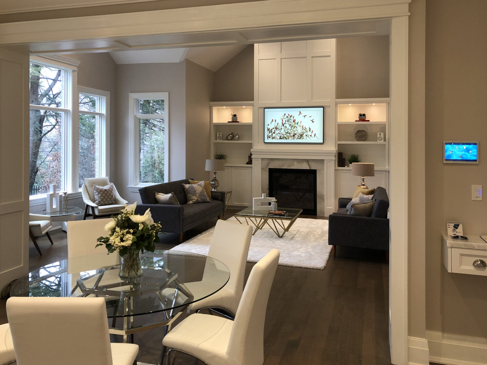 Spacious living room with modern decor, featuring a fireplace, glass dining table, cozy sofa seating, and large windows overlooking trees. Neutral color palette.