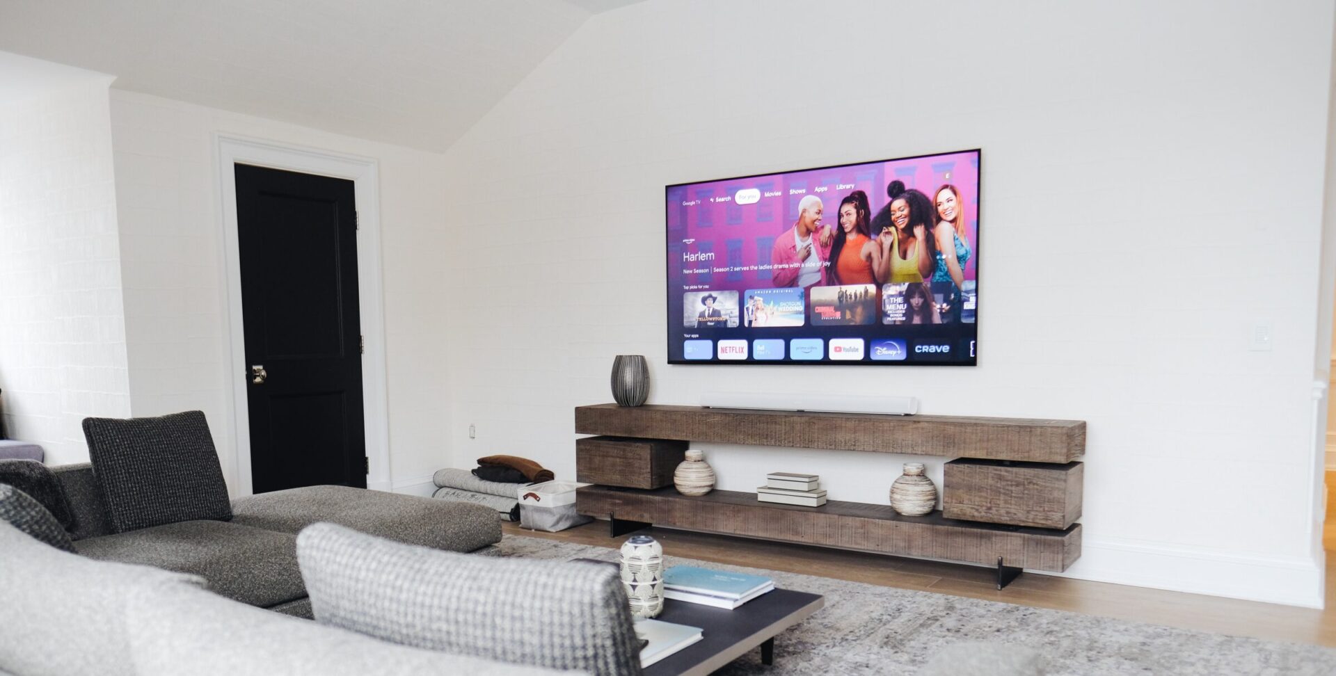 A modern living room features a wall-mounted TV displaying a streaming interface, with gray sofas and a wooden TV stand holding decorative items.