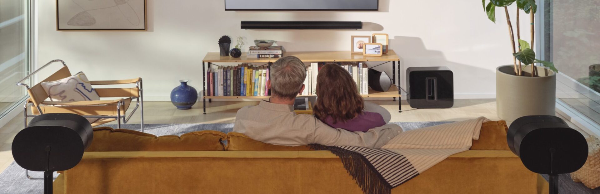 Two people sit on a sofa in a modern living room, watching TV. A bookshelf and plants decorate the cozy space.