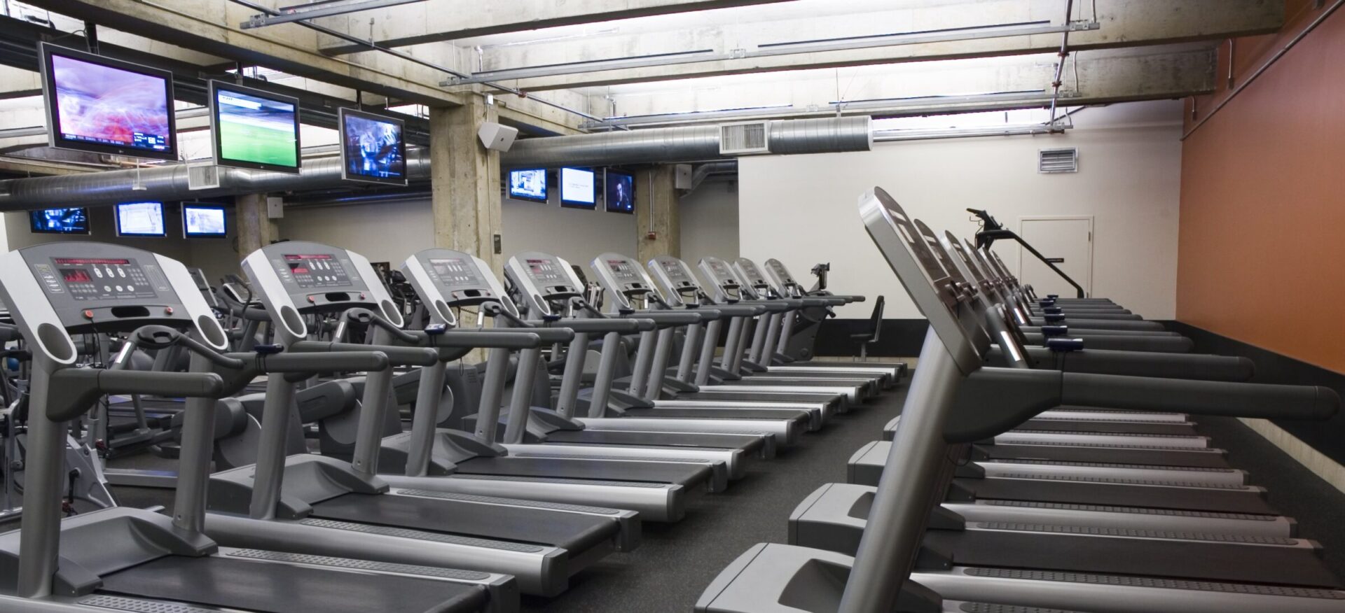 Rows of treadmills in a gym with multiple screens displaying various channels. The space is well-lit with an industrial-style ceiling.