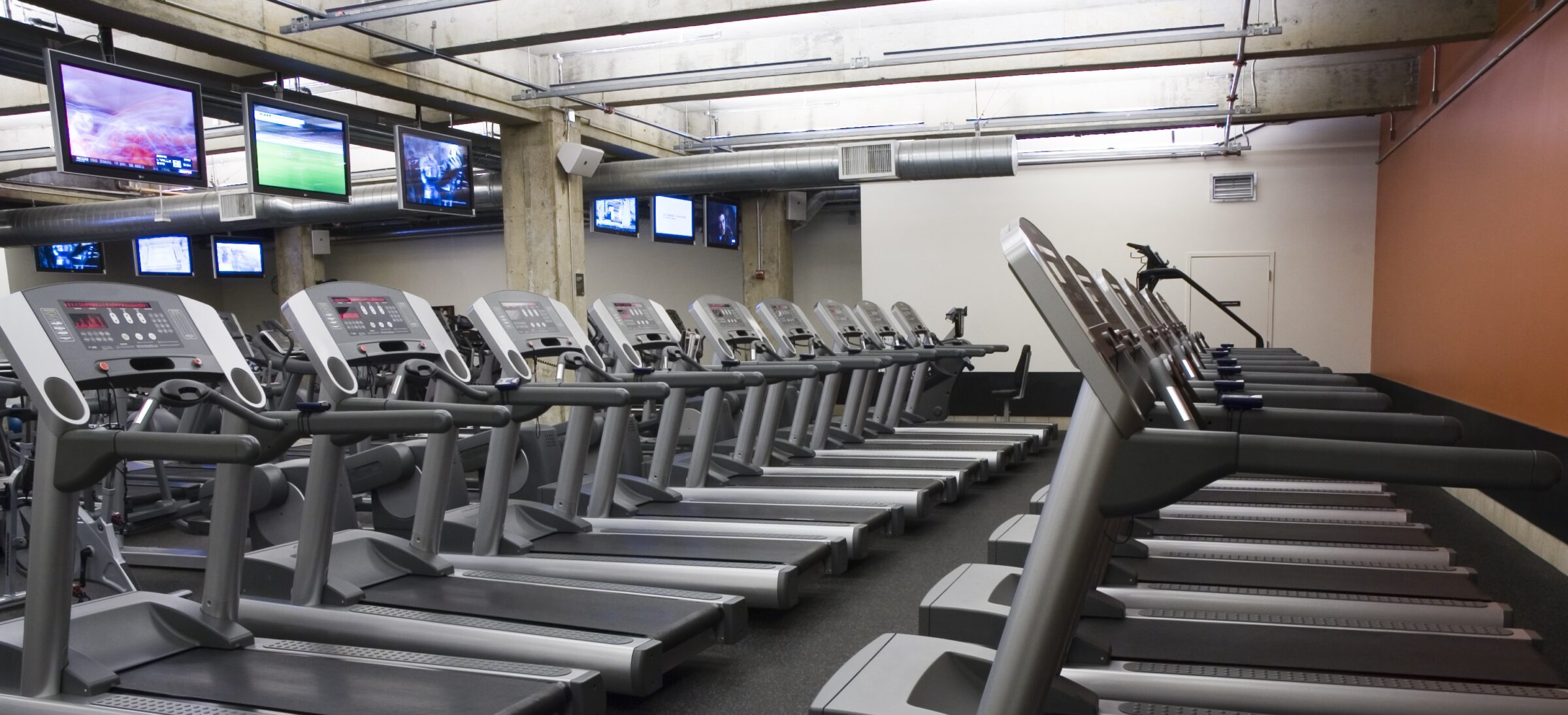 Rows of treadmills in a gym with multiple screens displaying various channels. The space is well-lit with an industrial-style ceiling.