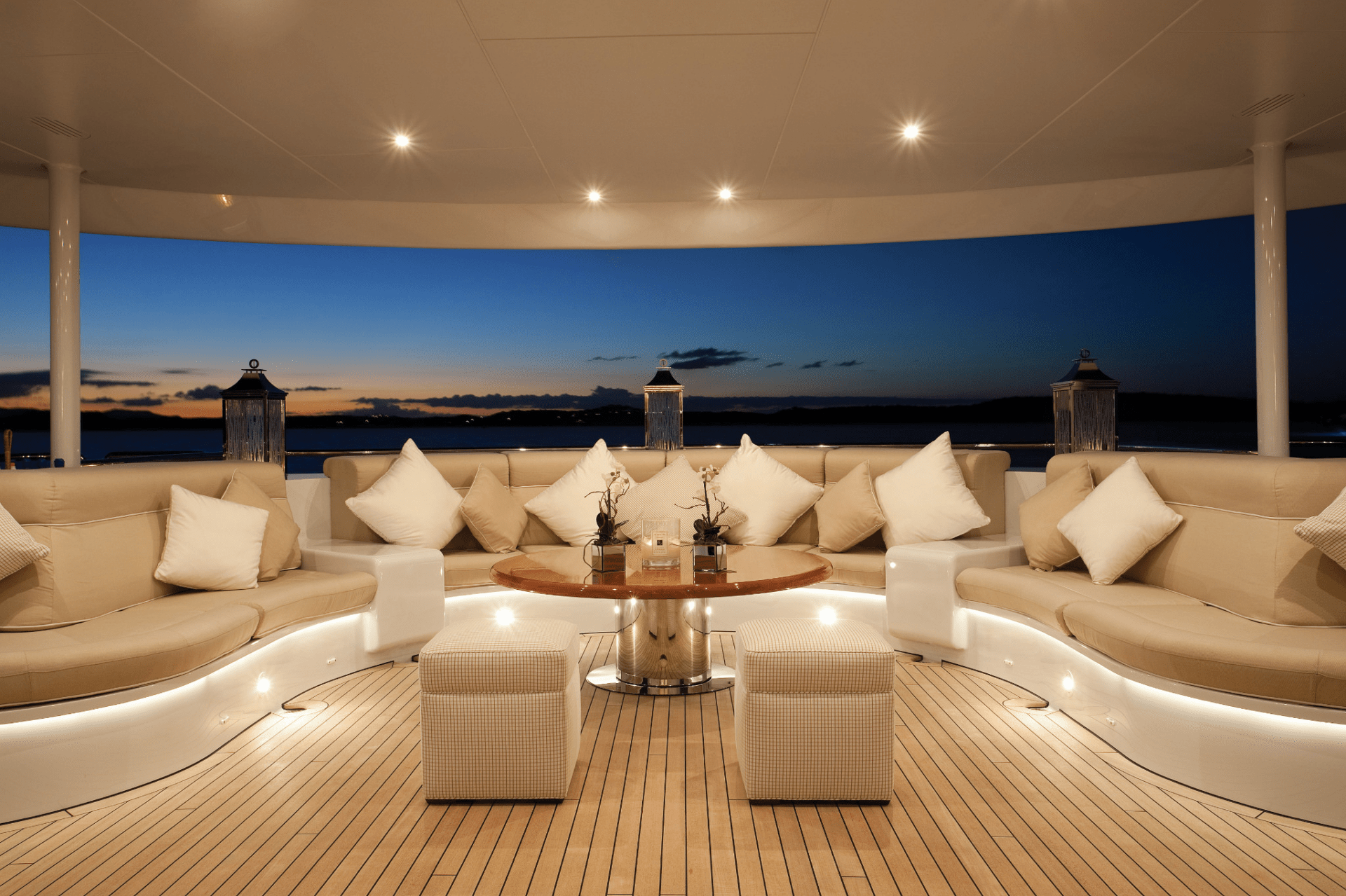 Luxurious yacht deck at sunset, featuring plush seating with cushions, ambient lighting, and panoramic ocean views. Lanterns enhance the tranquil atmosphere.