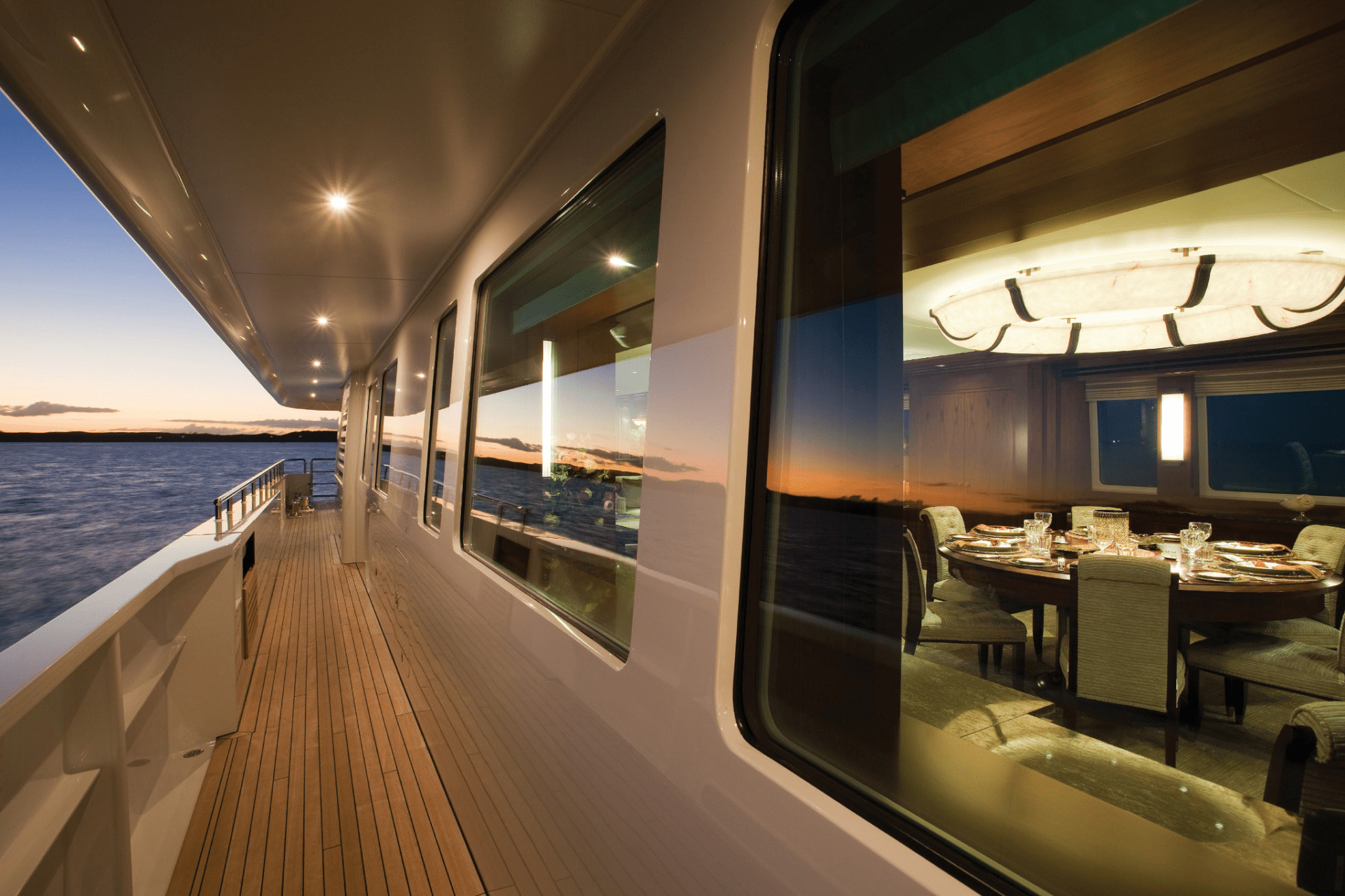 A yacht deck at sunset, overlooking the ocean. Inside, a dining table is elegantly set for an evening meal, reflecting warm light.