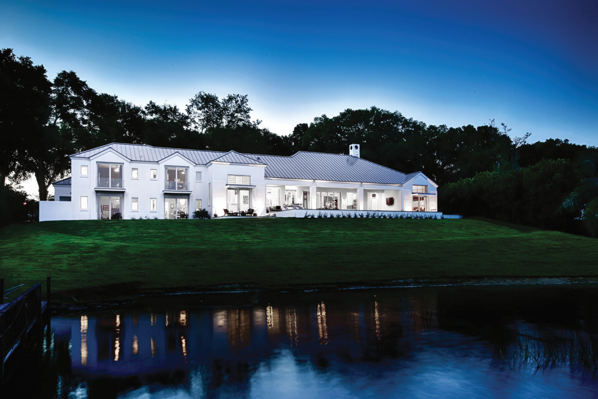 Expansive modern white house illuminated at dusk, set against lush greenery and a reflective pond, creating a serene and luxurious atmosphere.