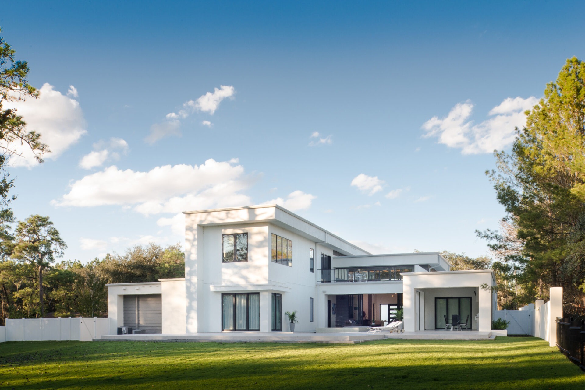 Modern white two-story house surrounded by trees, with large windows and a spacious lawn, under a clear blue sky with scattered clouds.