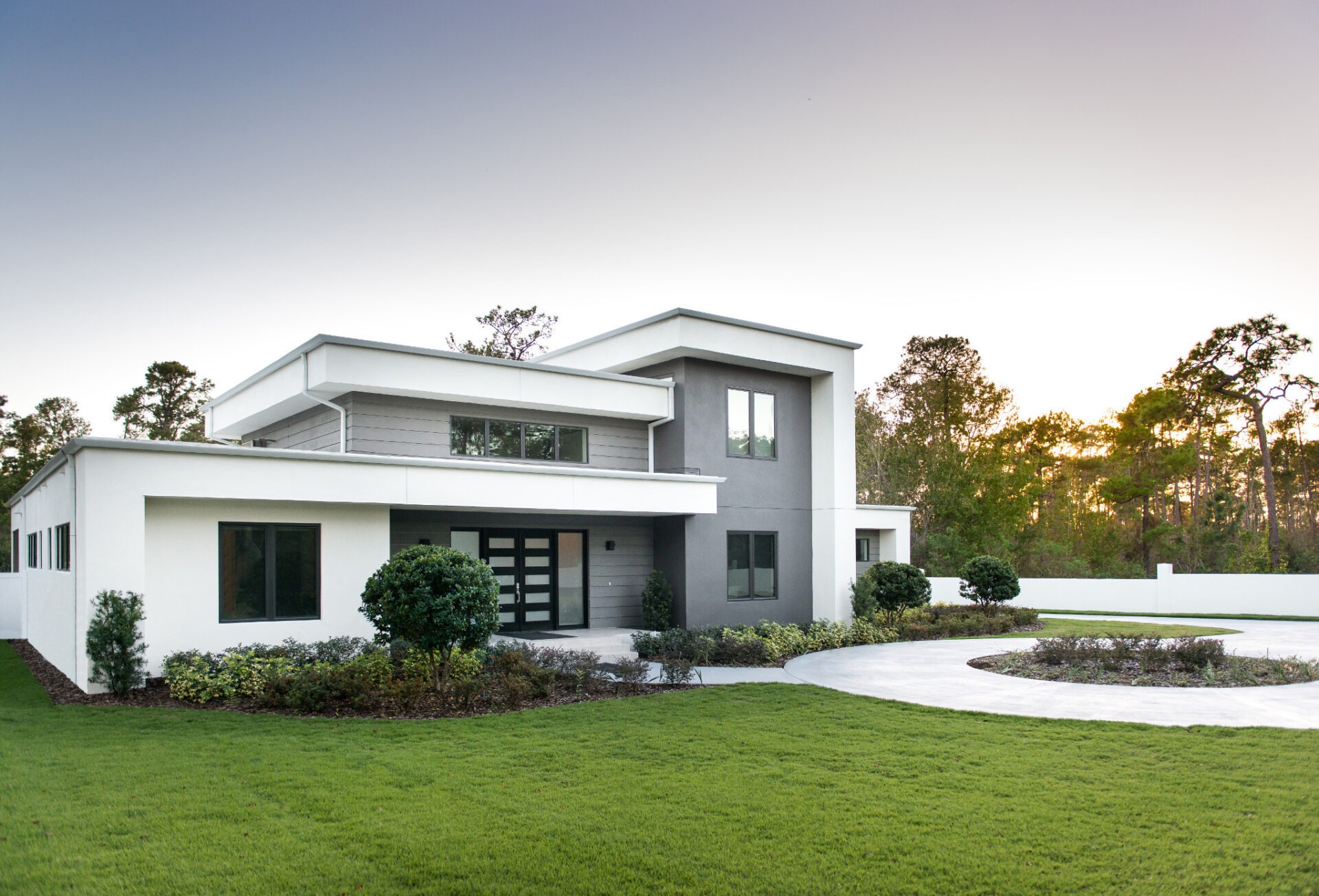 Modern two-story house with minimalist design, large windows, and a manicured lawn. Surrounded by trees, under a clear sky at dusk.