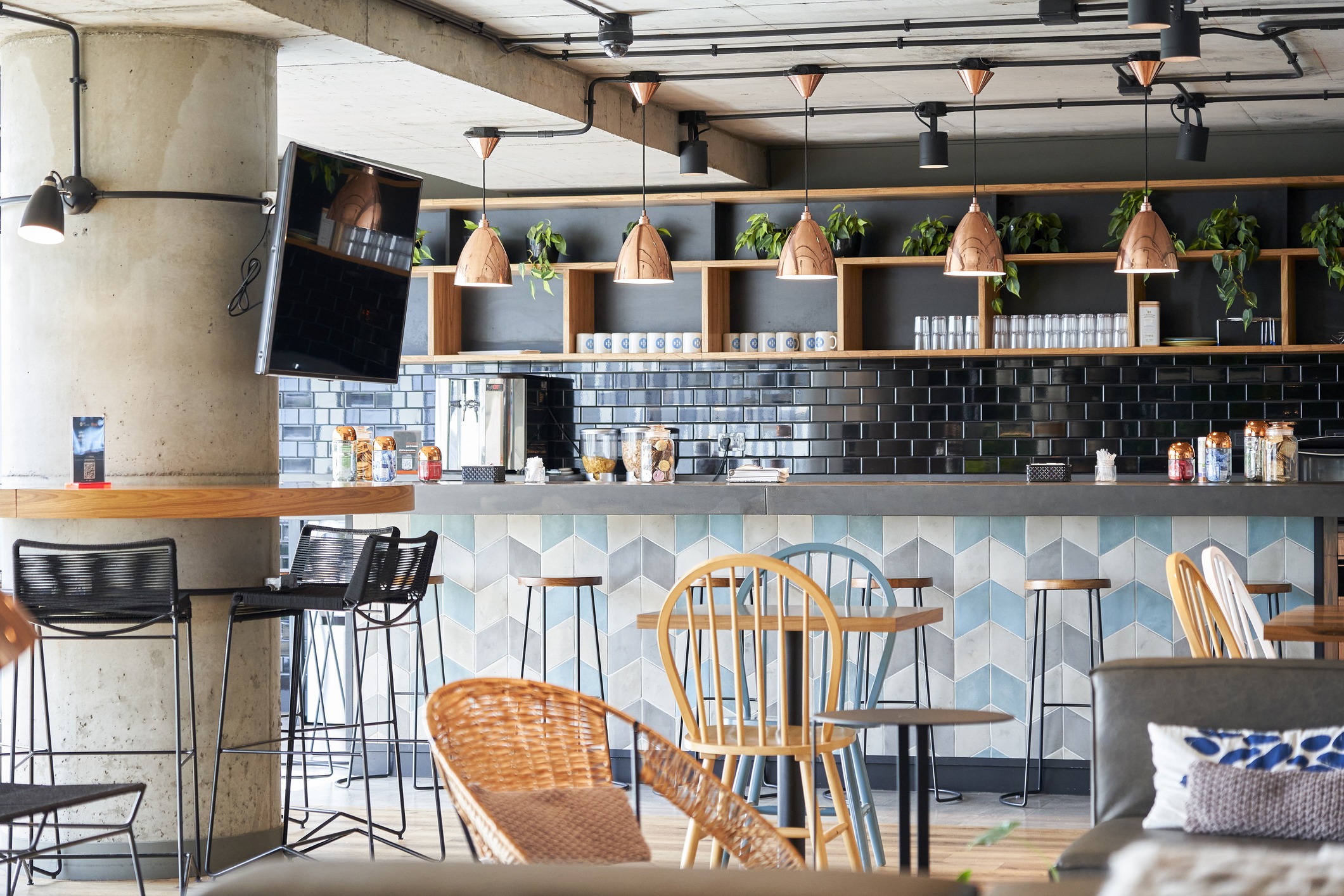 Modern café interior with geometric patterned bar front, hanging copper lamps, and green plants on shelves. Wooden and wicker chairs surround tables.