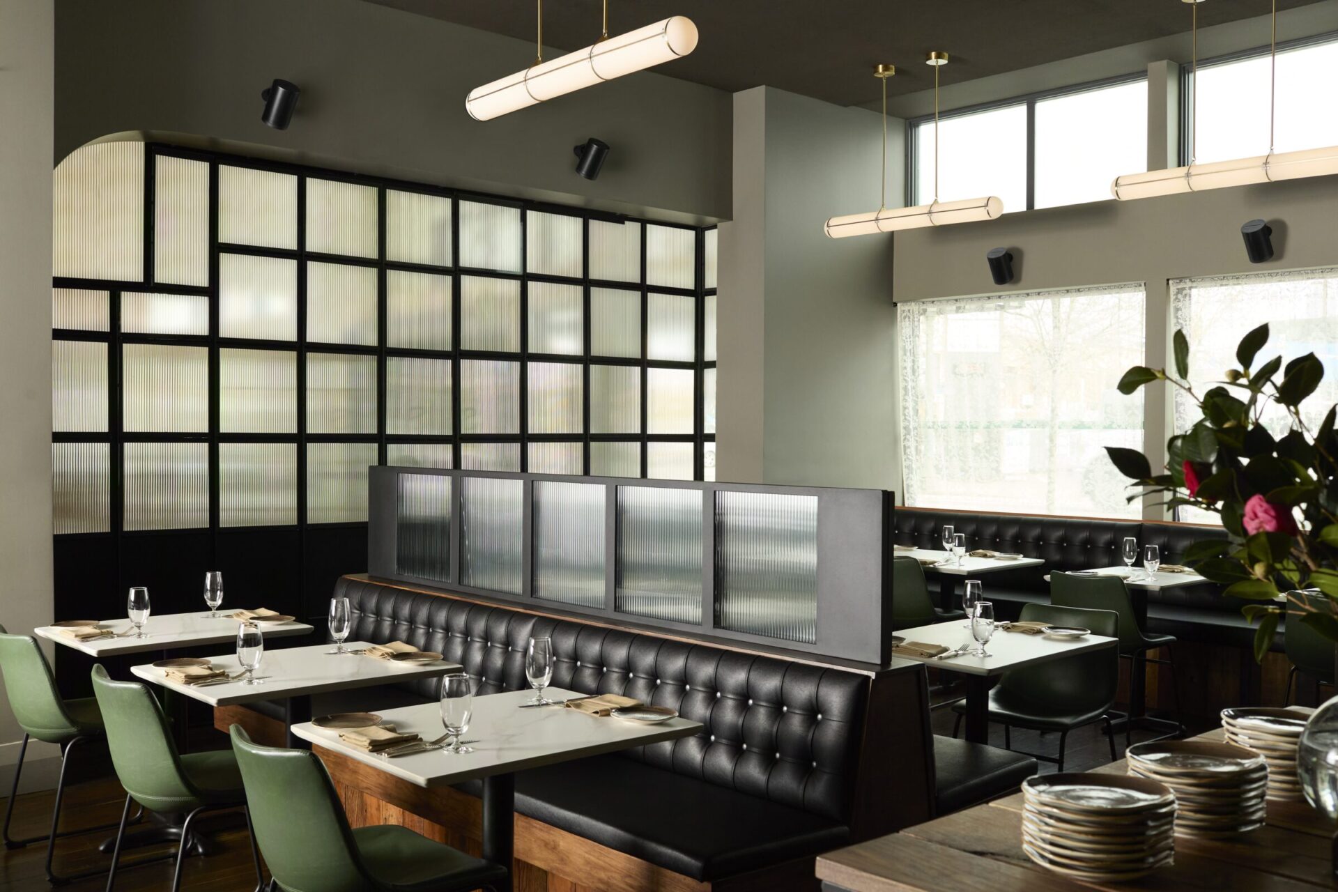 Modern restaurant interior with stylish black and green seating, frosted glass dividers, and elegant lighting. Tables set with glasses and plates.