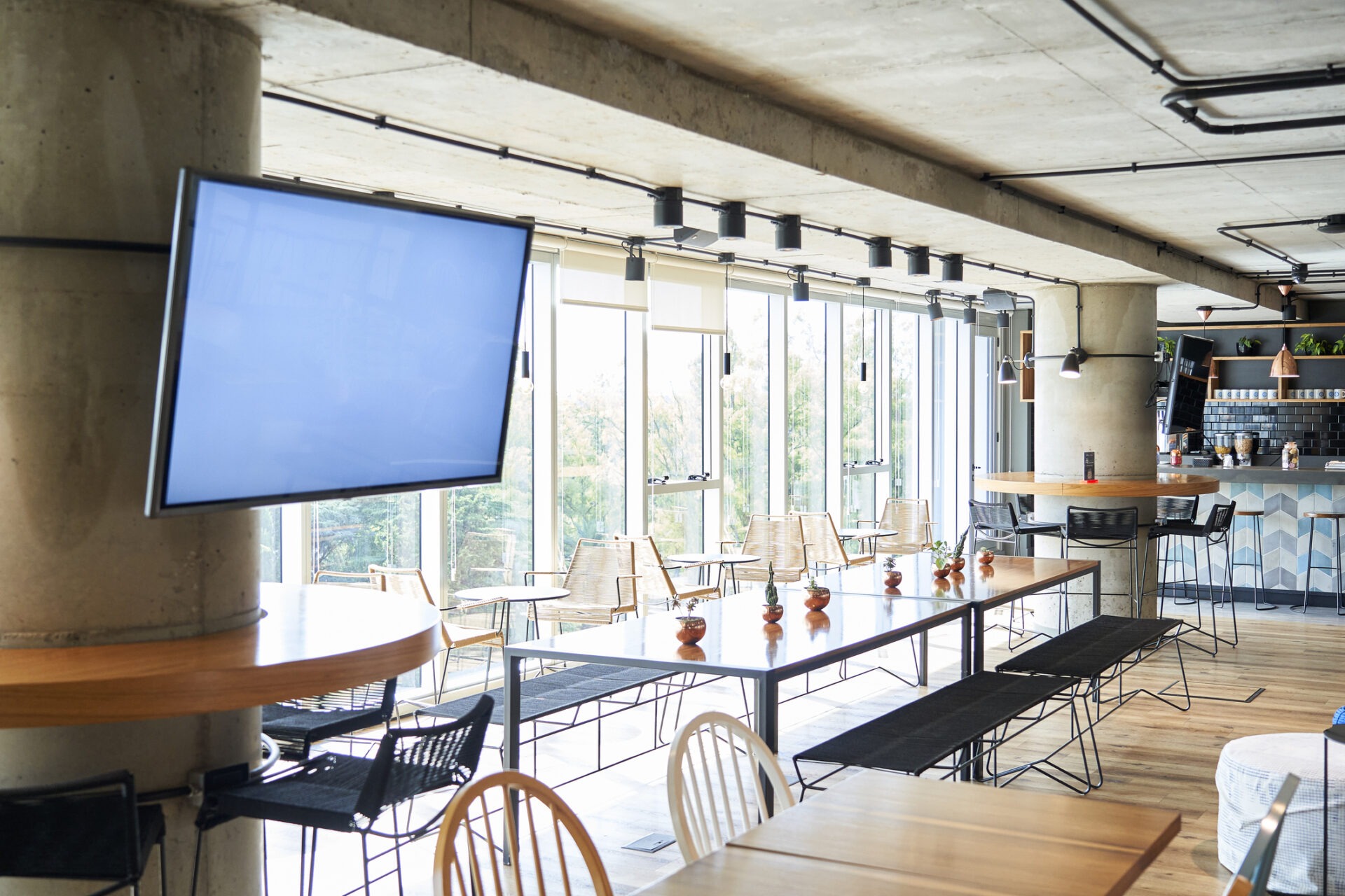 Modern café interior with large windows, wooden tables, and chairs. A television screen is mounted on a pillar. The atmosphere is bright and inviting.