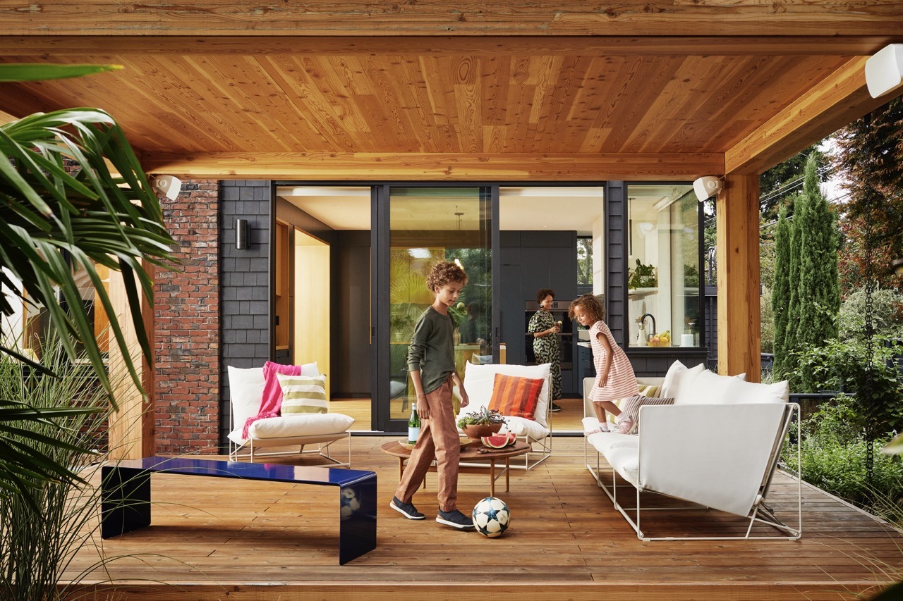 A modern patio with two children playing soccer, a person inside, and contemporary furniture. Wooden ceiling, glass doors, and lush greenery.