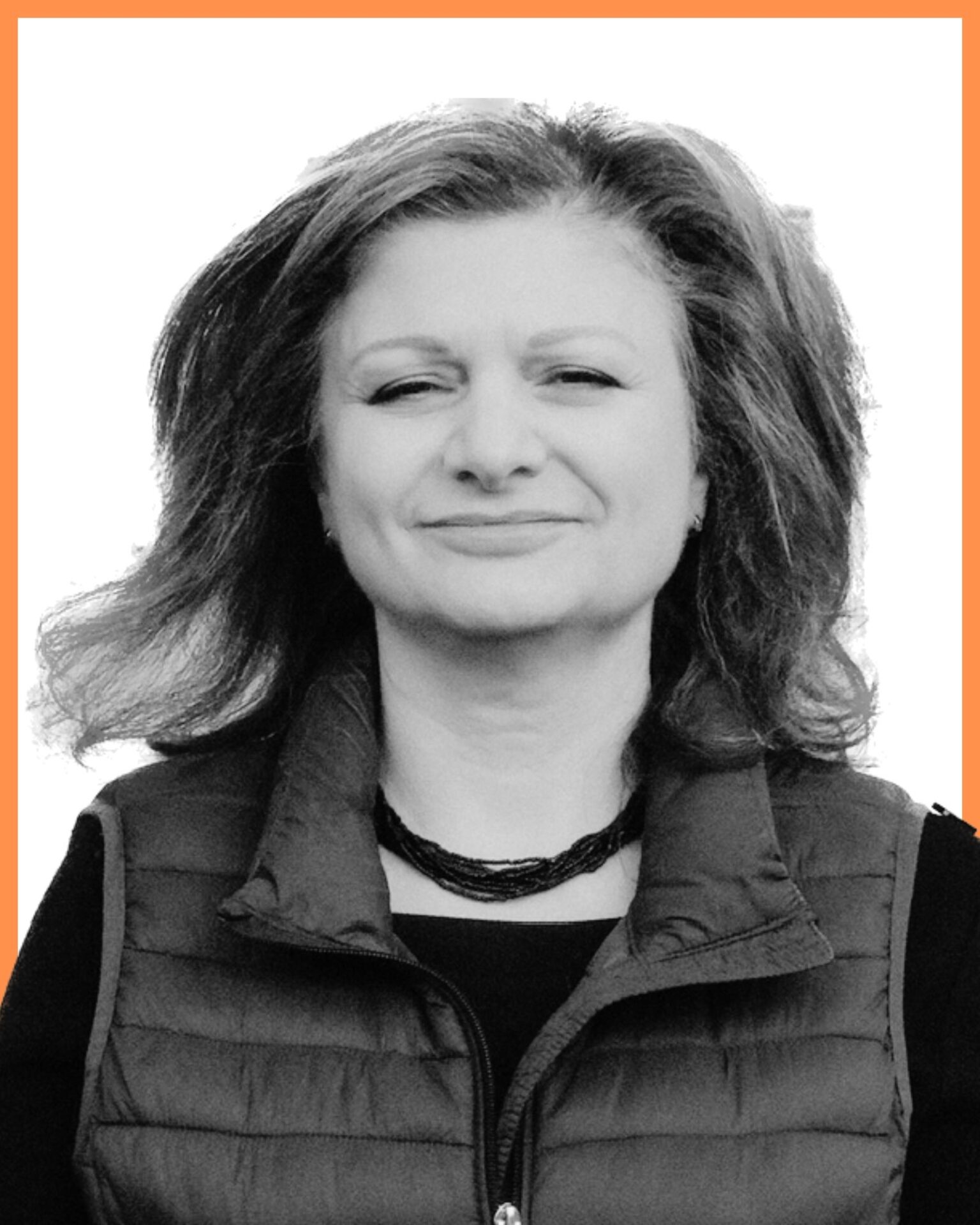 A person with medium-length hair wearing a dark puffer vest and necklace, set against a plain white background with an orange border.