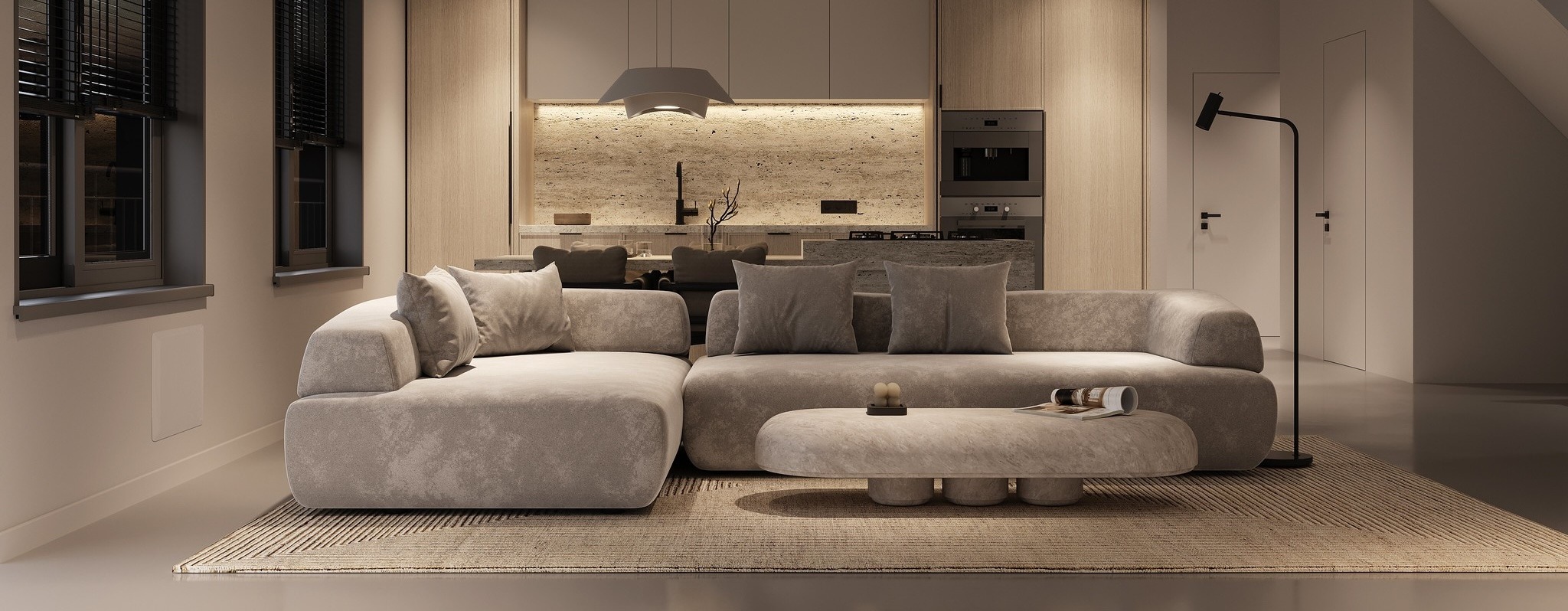 A modern living room with a large gray sectional sofa, a minimalist coffee table, and a built-in kitchen featuring warm lighting and wood accents.