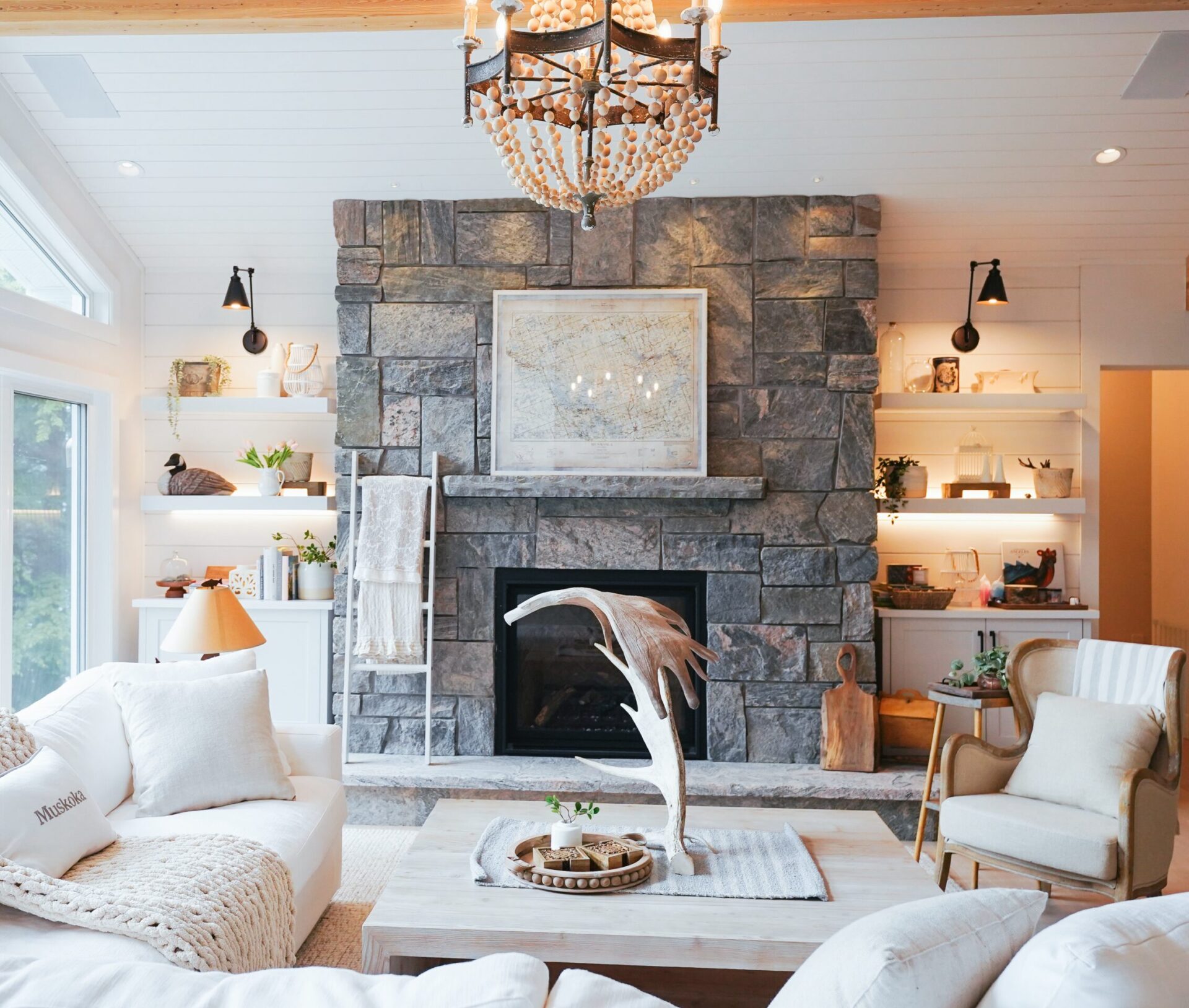 Cozy living room with stone fireplace, chandelier, white sofas, and decorative shelves. Large windows illuminate the rustic, elegant decor.