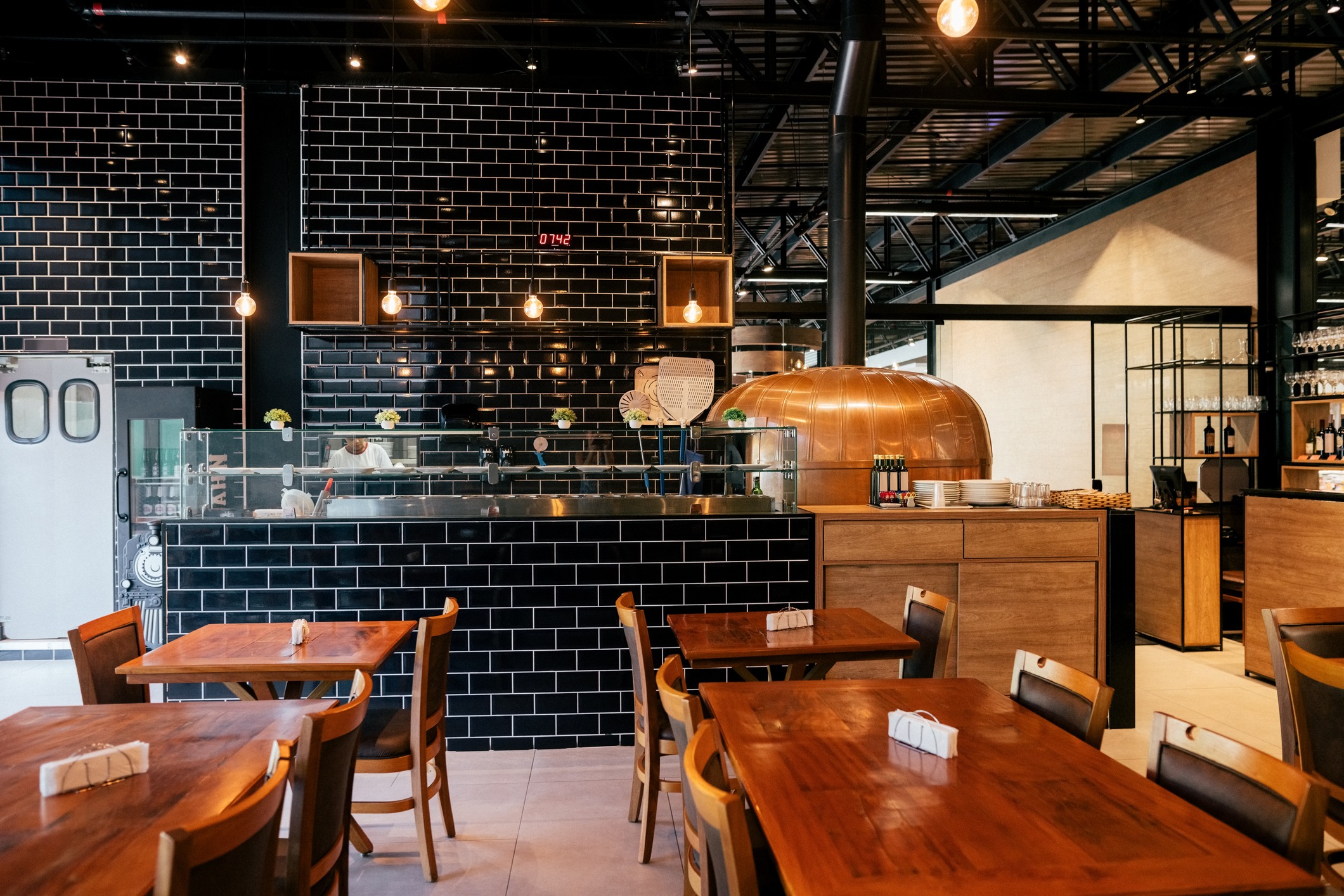 A cozy restaurant with wooden tables, black brick walls, and an open kitchen featuring a copper pizza oven. Warm lighting creates an inviting atmosphere.