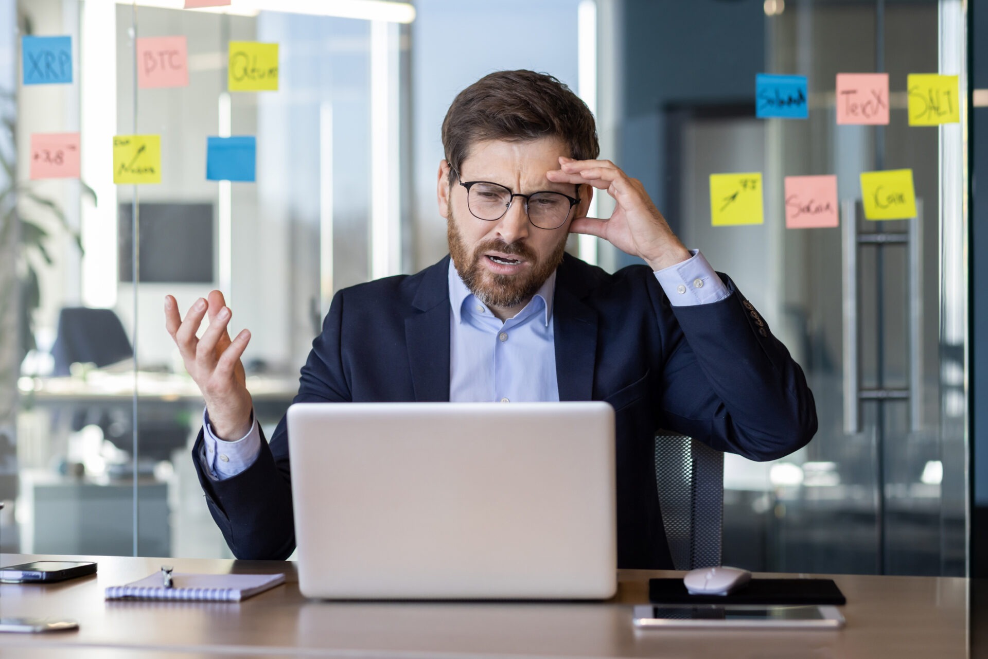 A person in a suit appears frustrated while looking at a laptop in an office with colorful sticky notes on a glass wall.