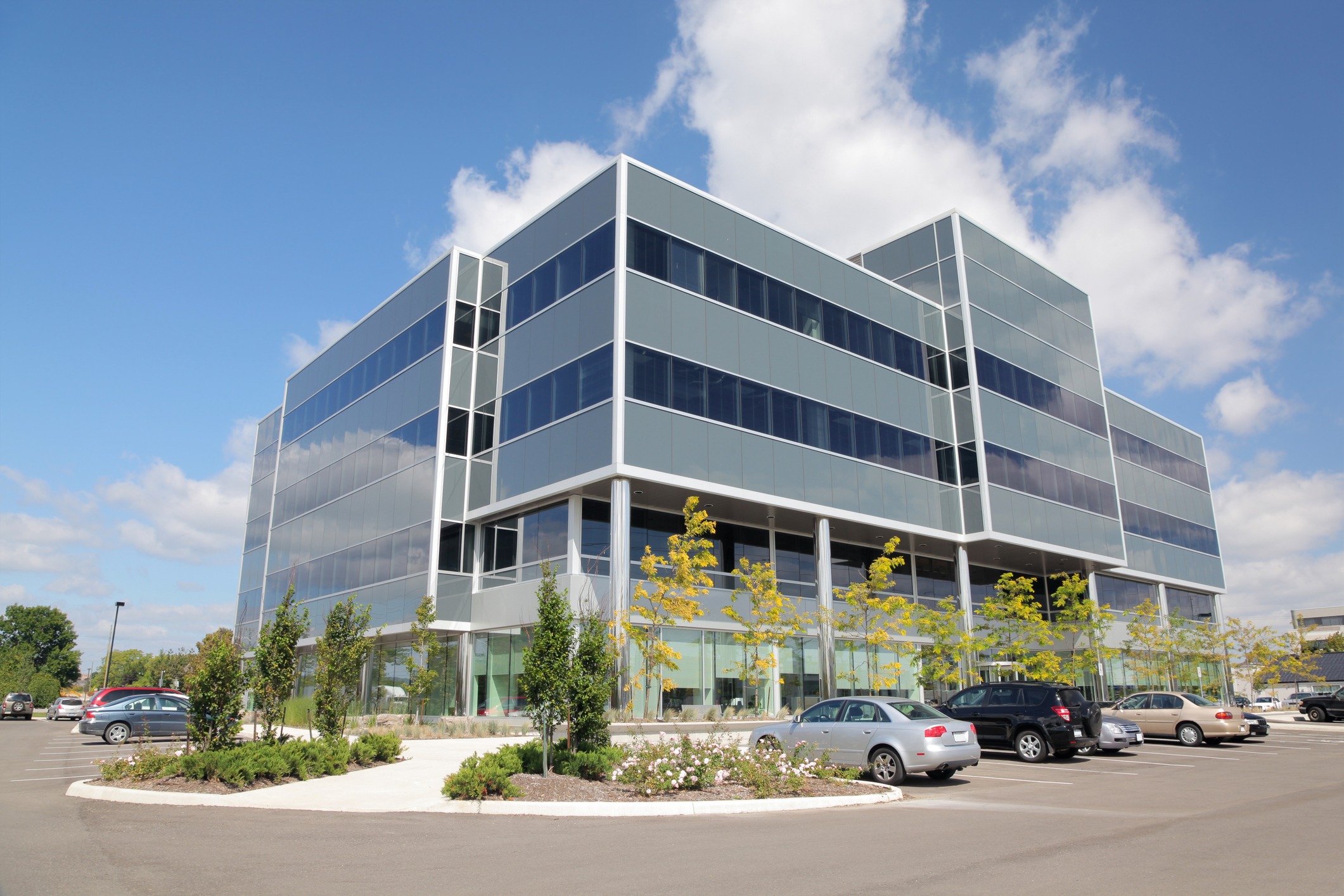 A modern office building with mirror-like glass exterior, surrounded by a parking lot. Several cars and greenery complement the clear, sunny day.
