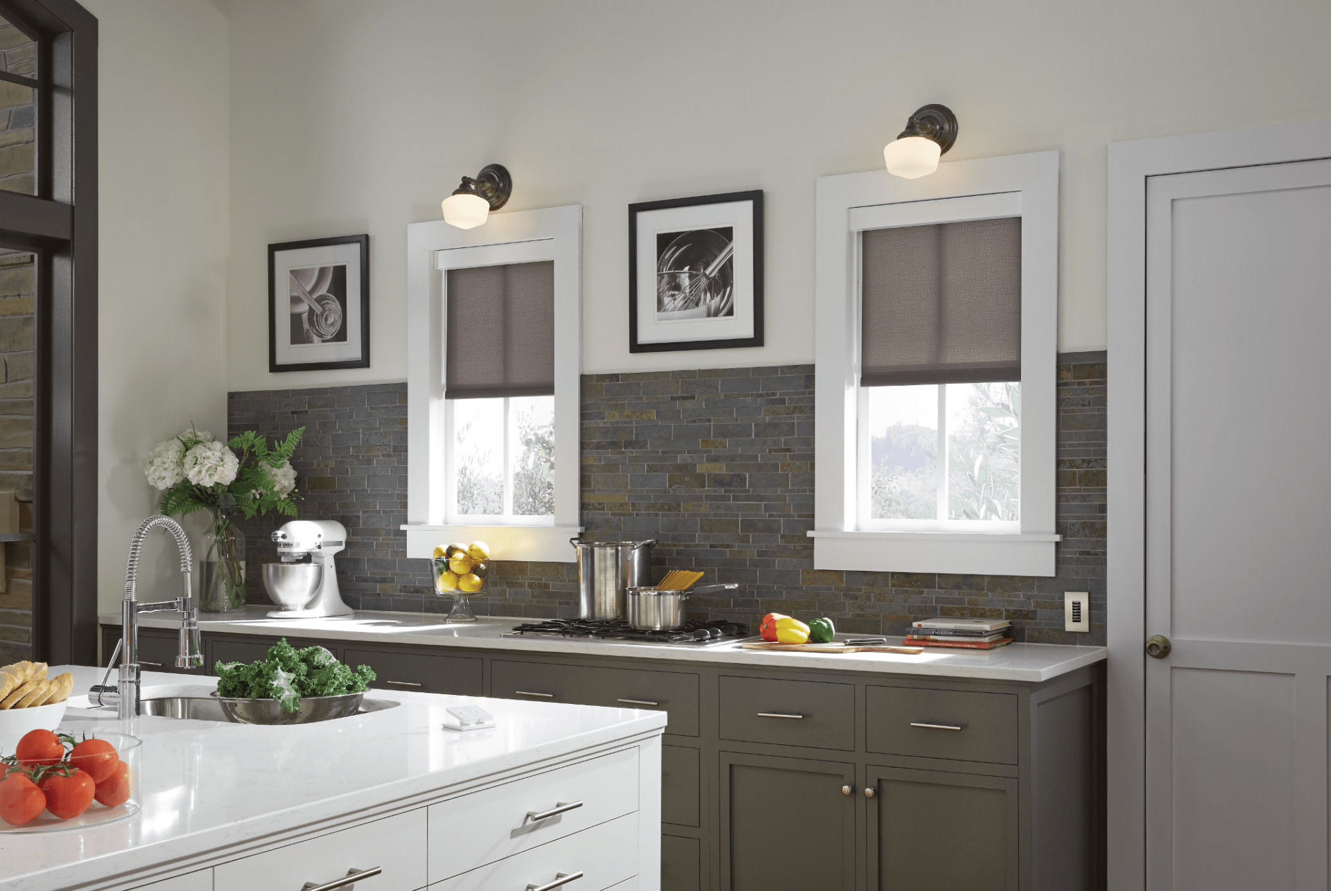 Modern kitchen with white cabinets, dark tiled backsplash, and stainless steel appliances. Countertop displays fresh produce. Two windows with gray blinds and framed photos.