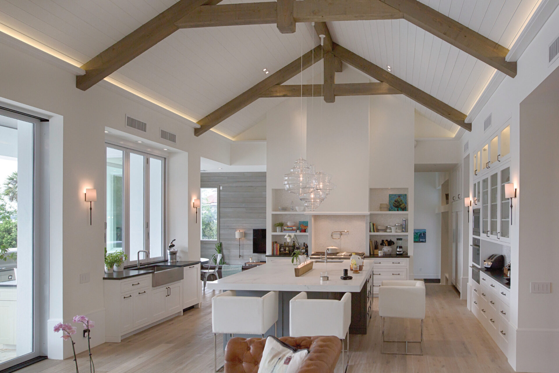 Spacious modern kitchen with vaulted wooden beams, large island with chairs, and bright lighting. White cabinets, wooden floors, and glass doors create an airy atmosphere.