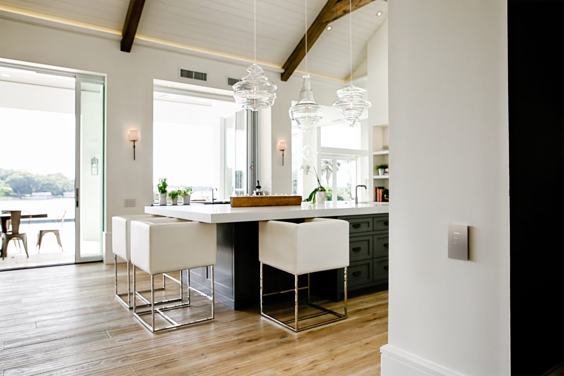 Modern kitchen with wooden floors, high ceilings, and large windows. Features white bar stools, hanging lights, and opens to a patio with a view.