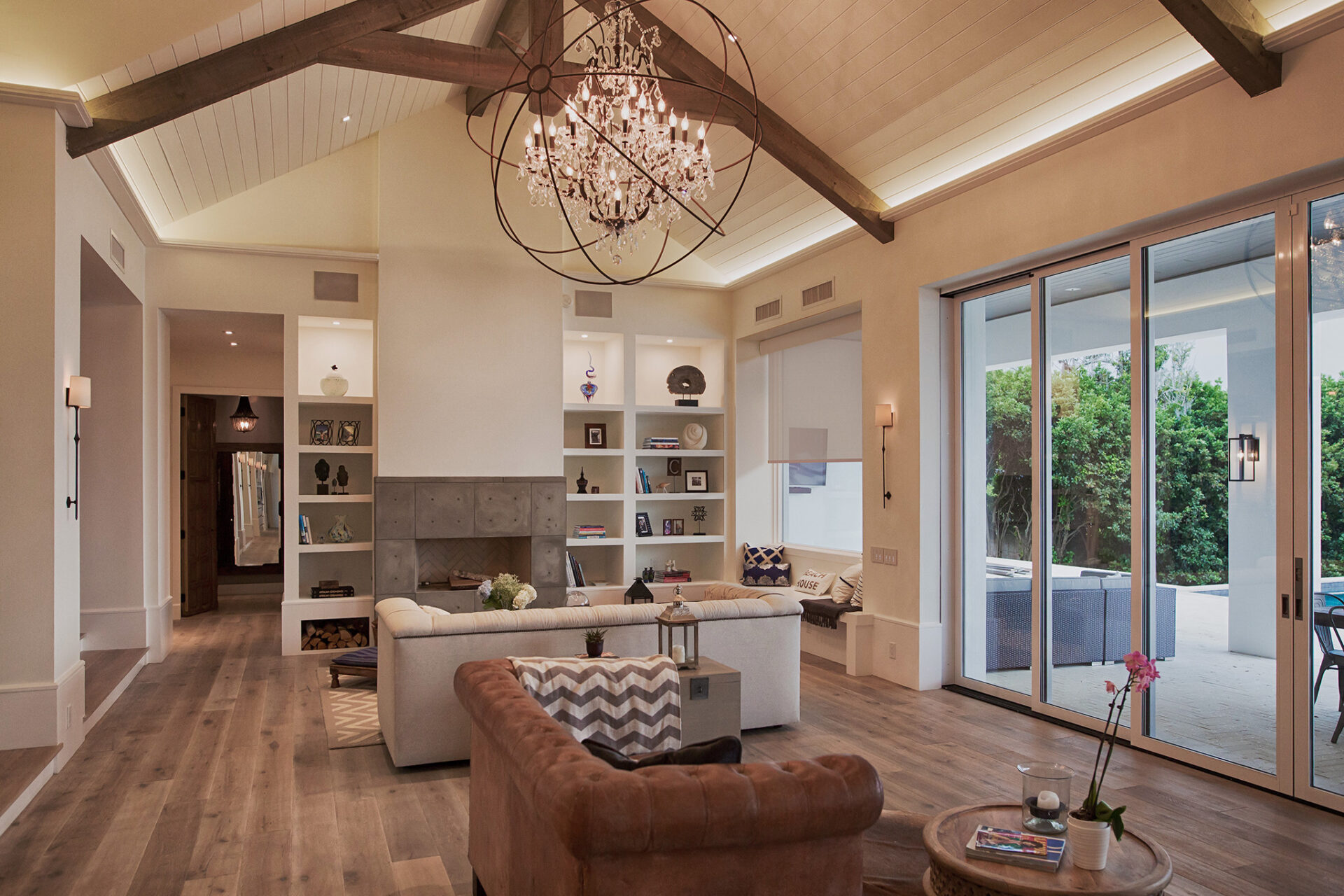 Spacious living room with modern decor, chandelier, wooden beams, and large sliding doors opening to outdoor greenery. Cozy seating area and decorative shelves.